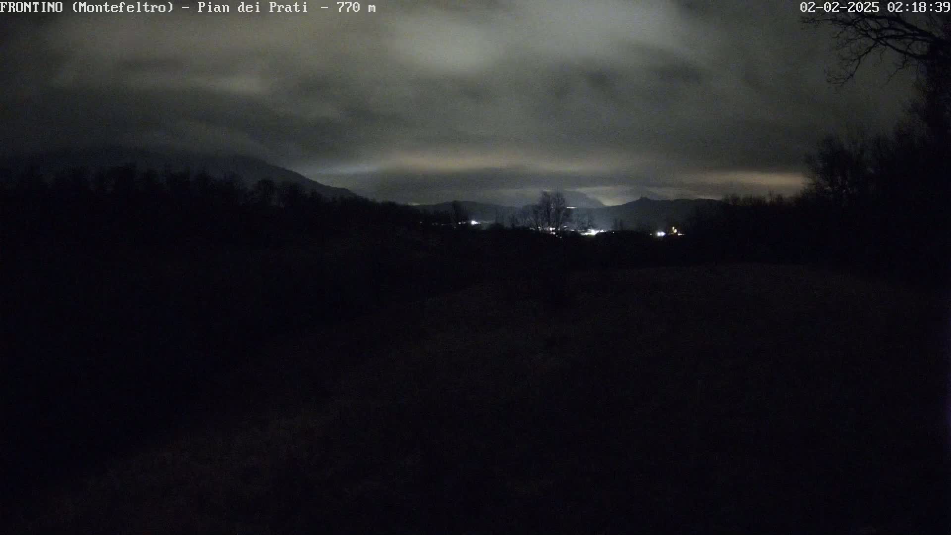 A dark, nighttime landscape shows distant lights in a valley below a cloudy sky.
