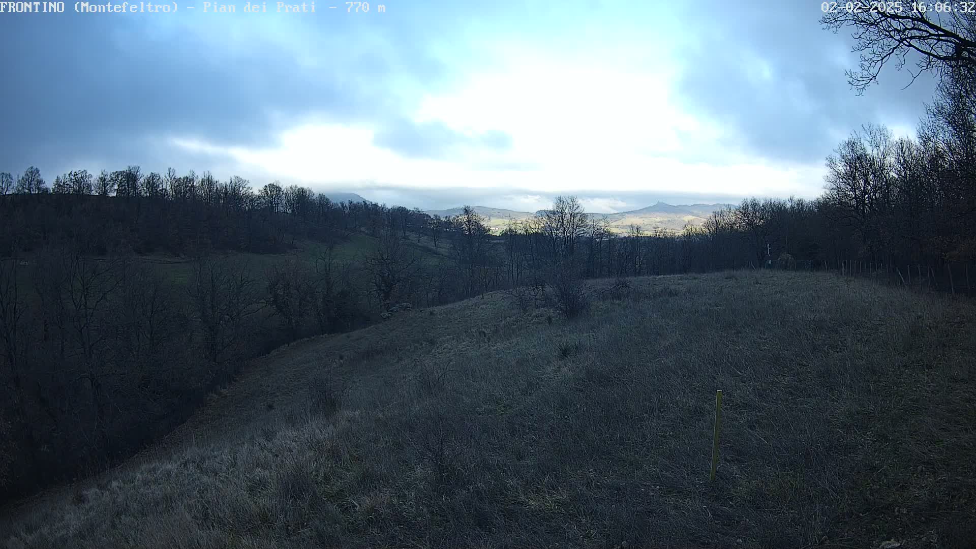 A partly cloudy day reveals a hilly landscape with leafless trees and brown, grassy fields stretching towards a distant mountain range under a mostly clear sky.