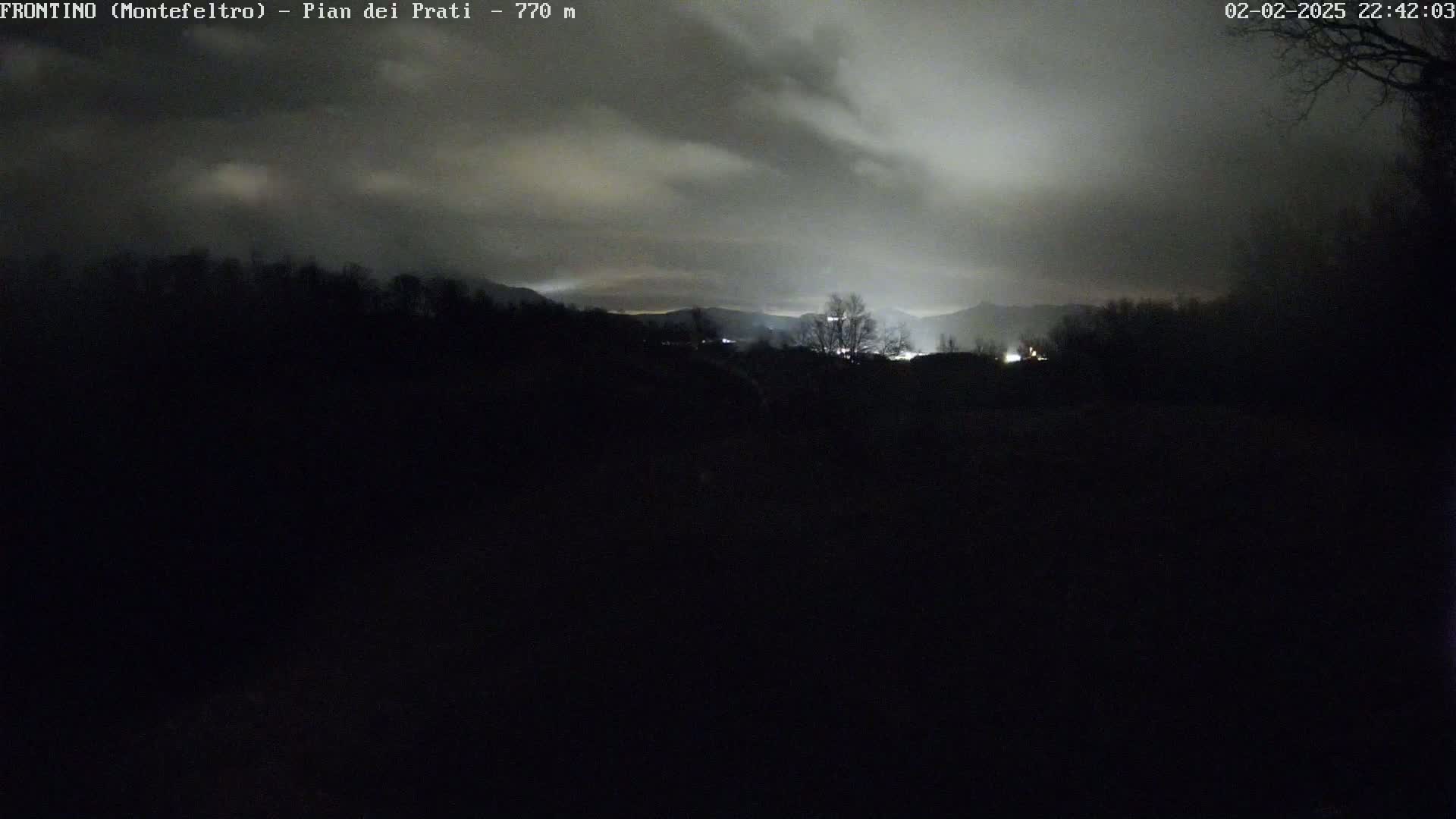 A nighttime view of a dark, silhouetted landscape under a cloudy sky, with distant lights visible in the background.