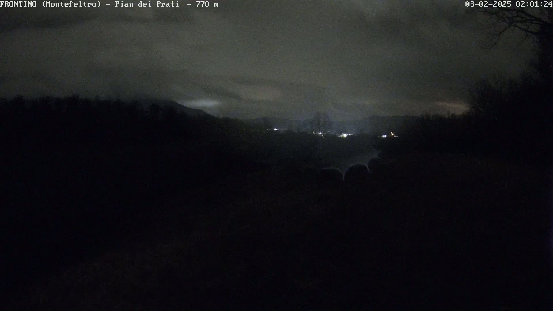 A nighttime view of a dark, hilly landscape with a few faint lights visible in the distance under a cloudy sky.