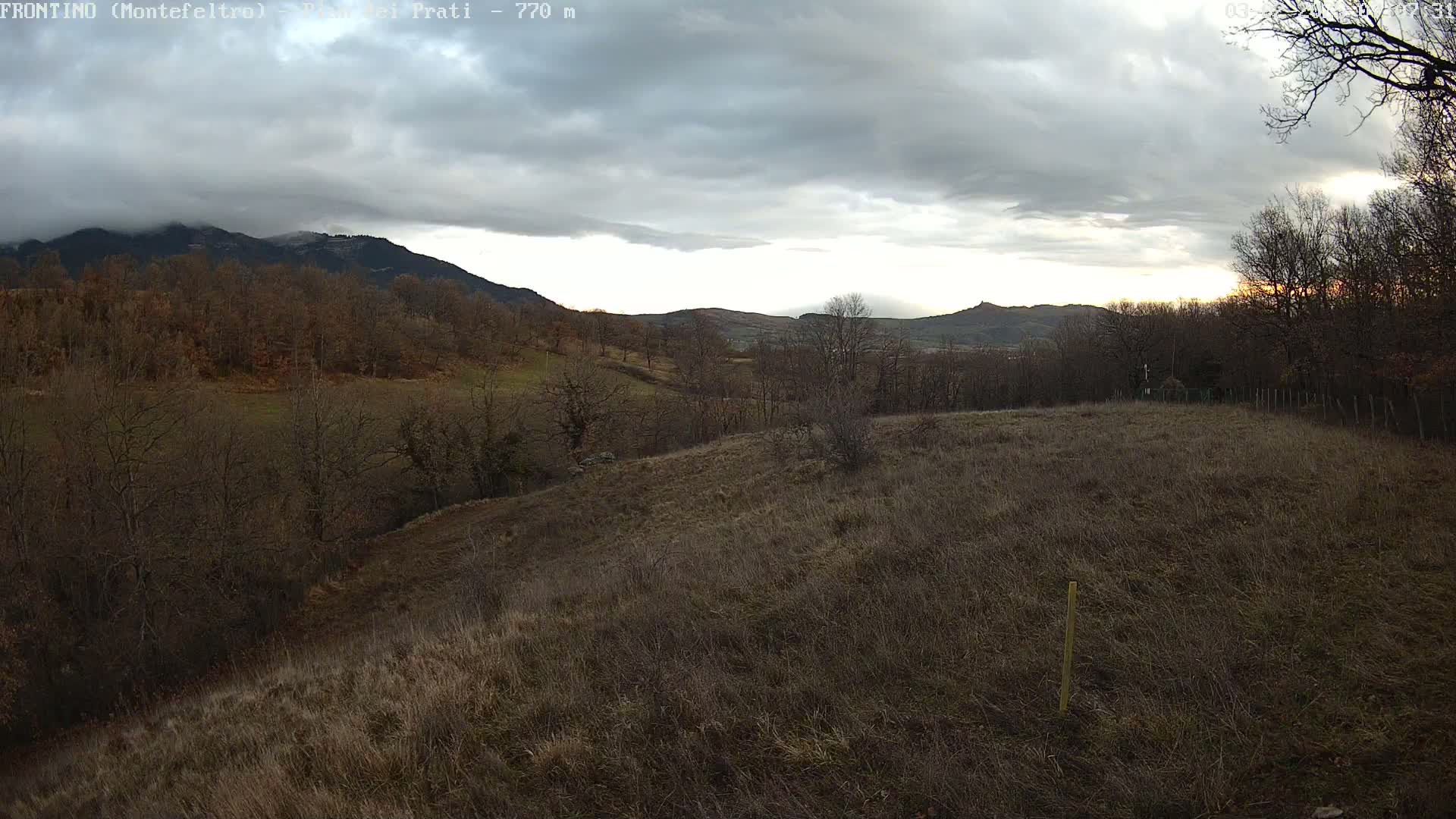 A mostly cloudy sky hangs over a hilly landscape of bare, brown fields and leafless trees, with distant mountains in the background.