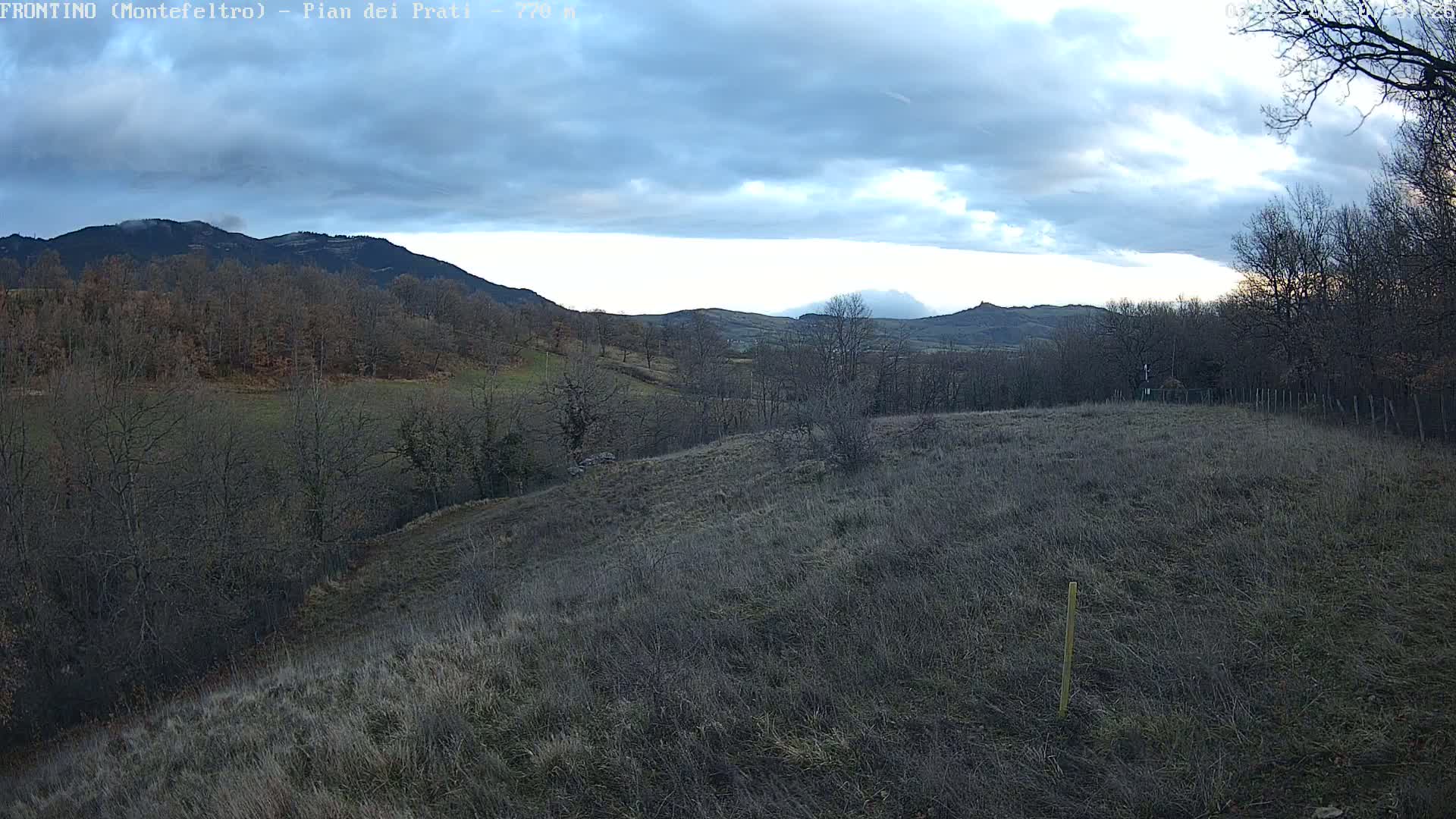 A view of a hilly landscape under a mostly cloudy sky, showing bare winter trees and dry, brown grass.