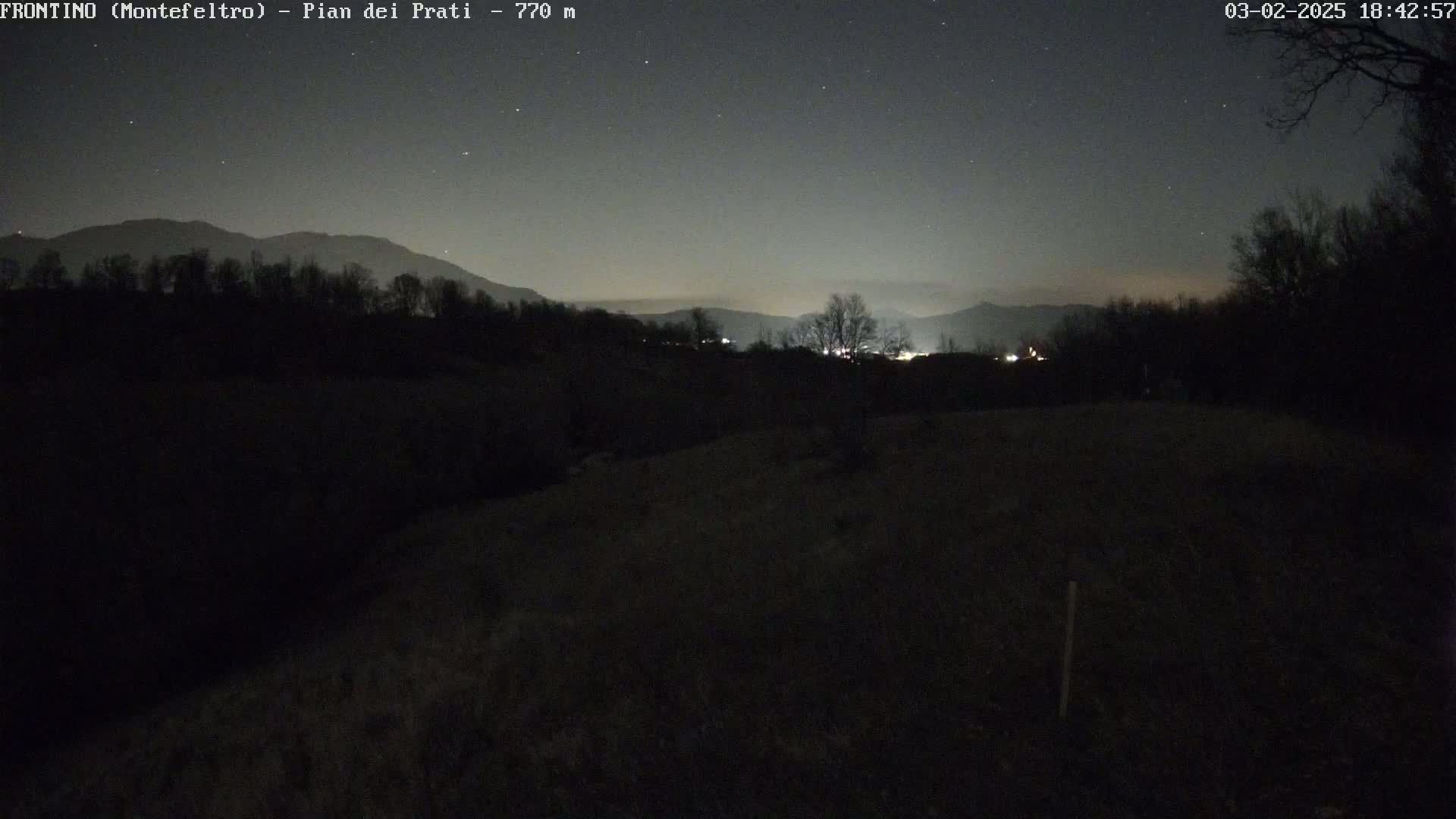 A nighttime view of a dark, hilly landscape under a clear, starlit sky shows a distant line of lights and silhouetted trees.