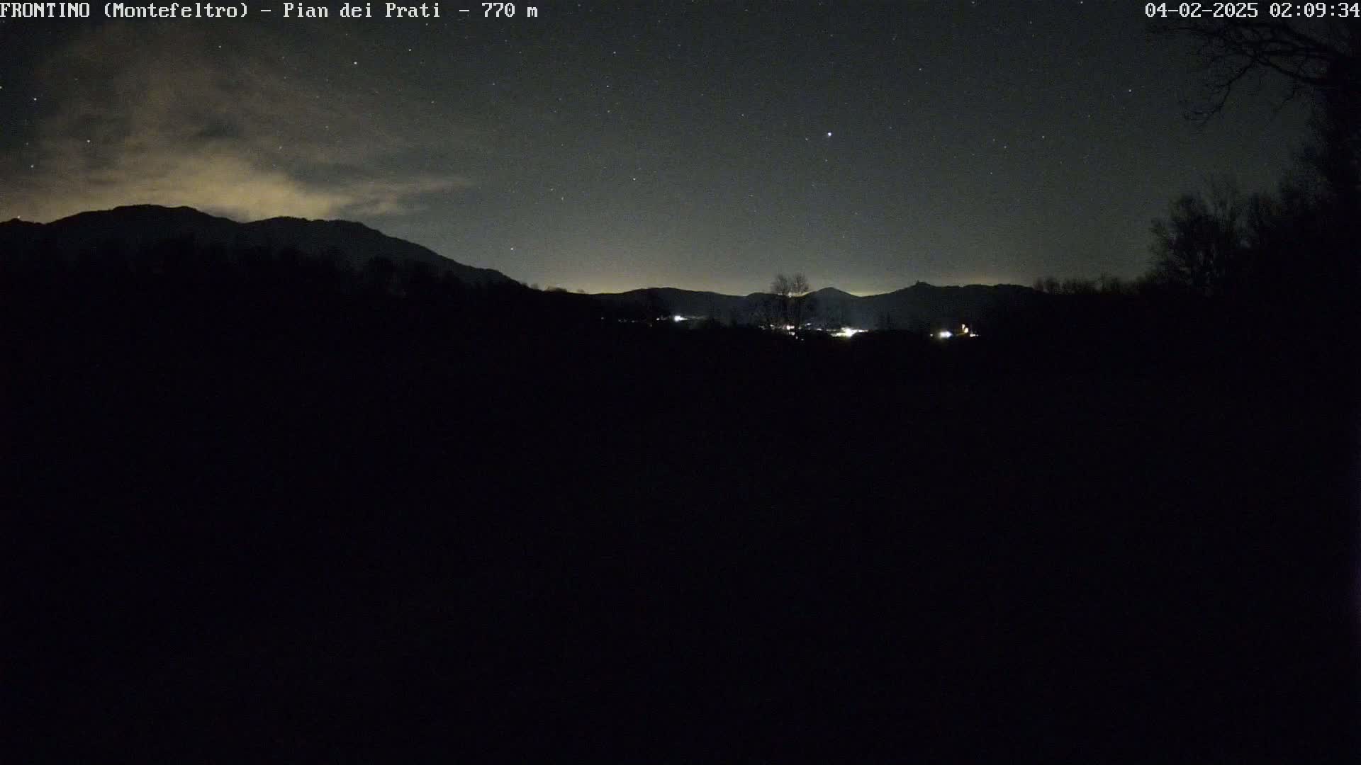 A nighttime view of a dark landscape with silhouetted mountains under a partly cloudy sky dotted with stars.