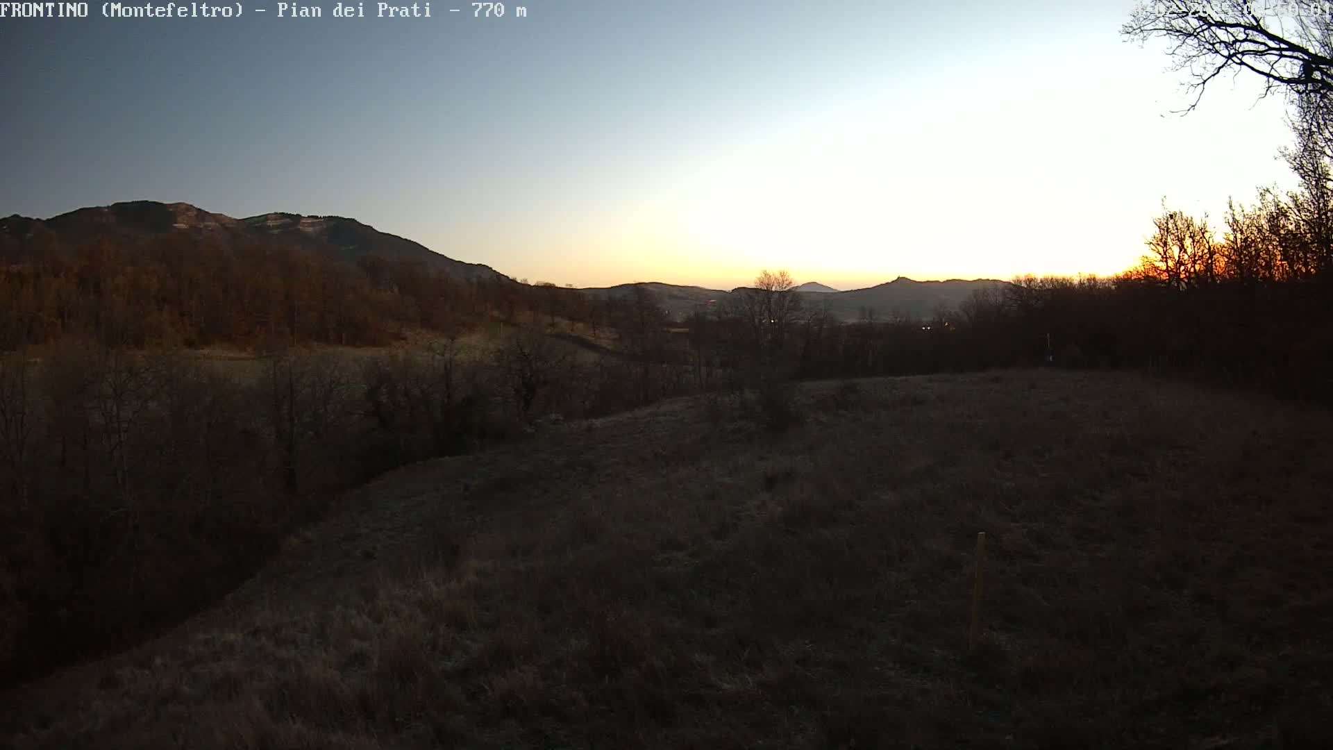 A sunset casts a warm glow over a hilly landscape of bare winter trees and brown fields.