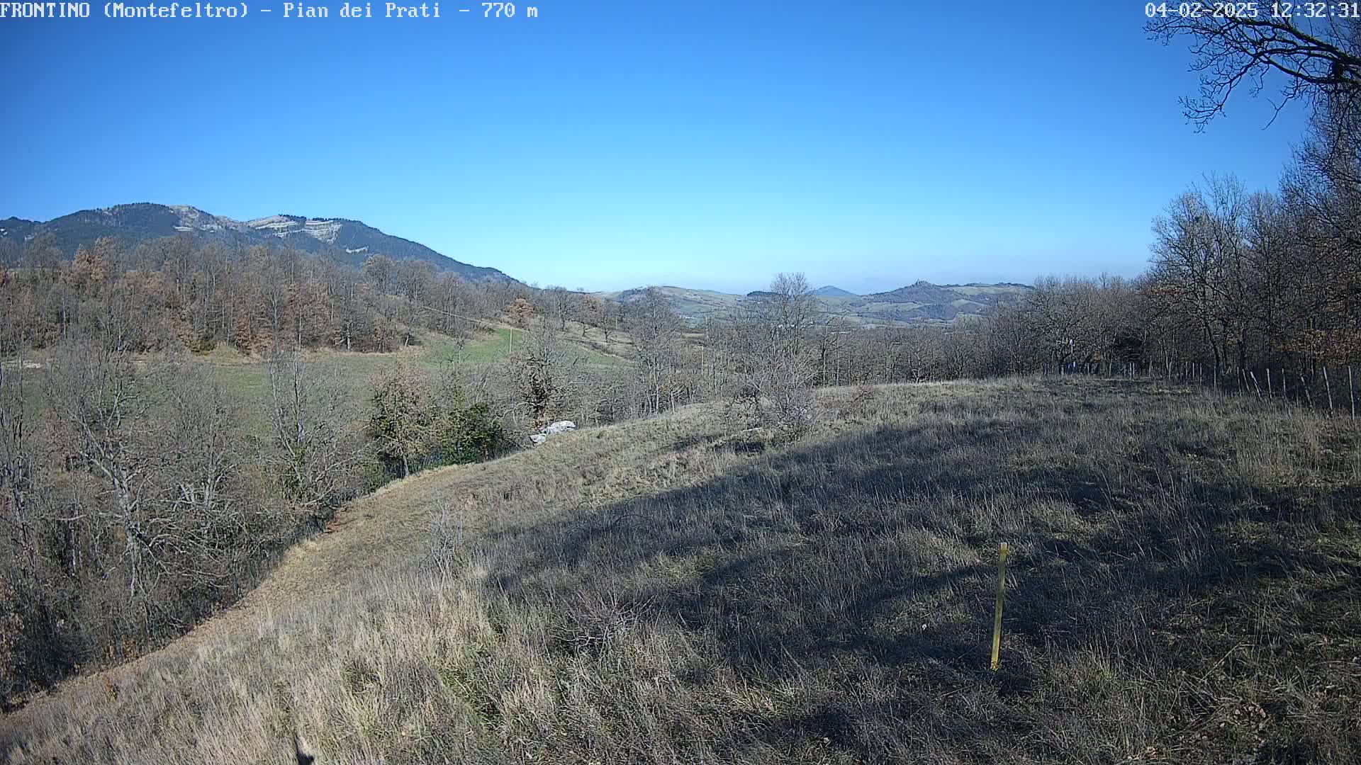 A sunny, clear day reveals a rolling landscape of dry, brown fields and leafless trees, with mountains in the background.