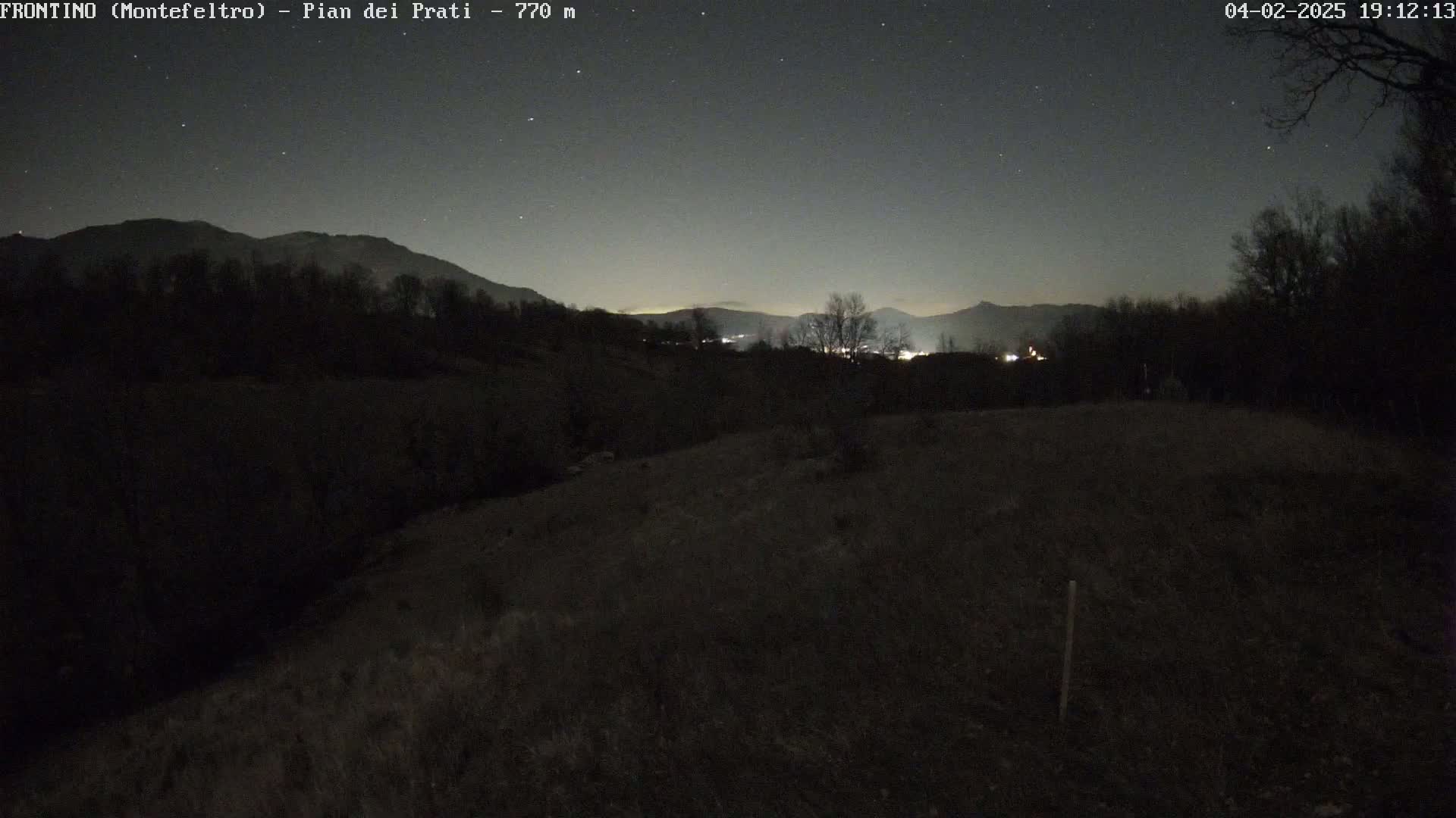 A nighttime view of a dark, grassy hillside under a clear, starry sky, with distant mountains and faint lights visible in the background.