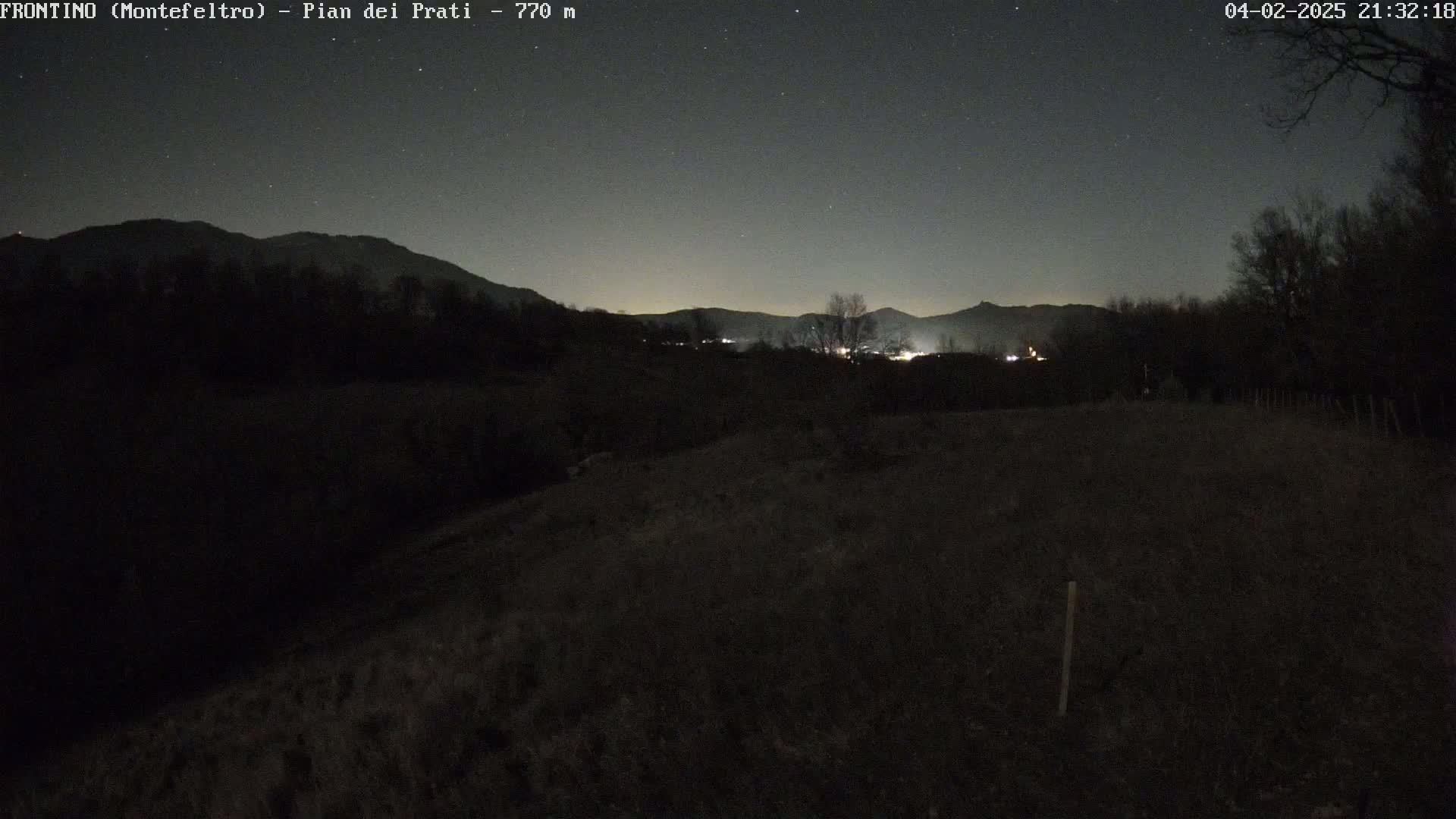 A nighttime, outdoor view of a dark, grassy field with a distant, dimly lit town nestled amongst dark hills under a starlit sky.