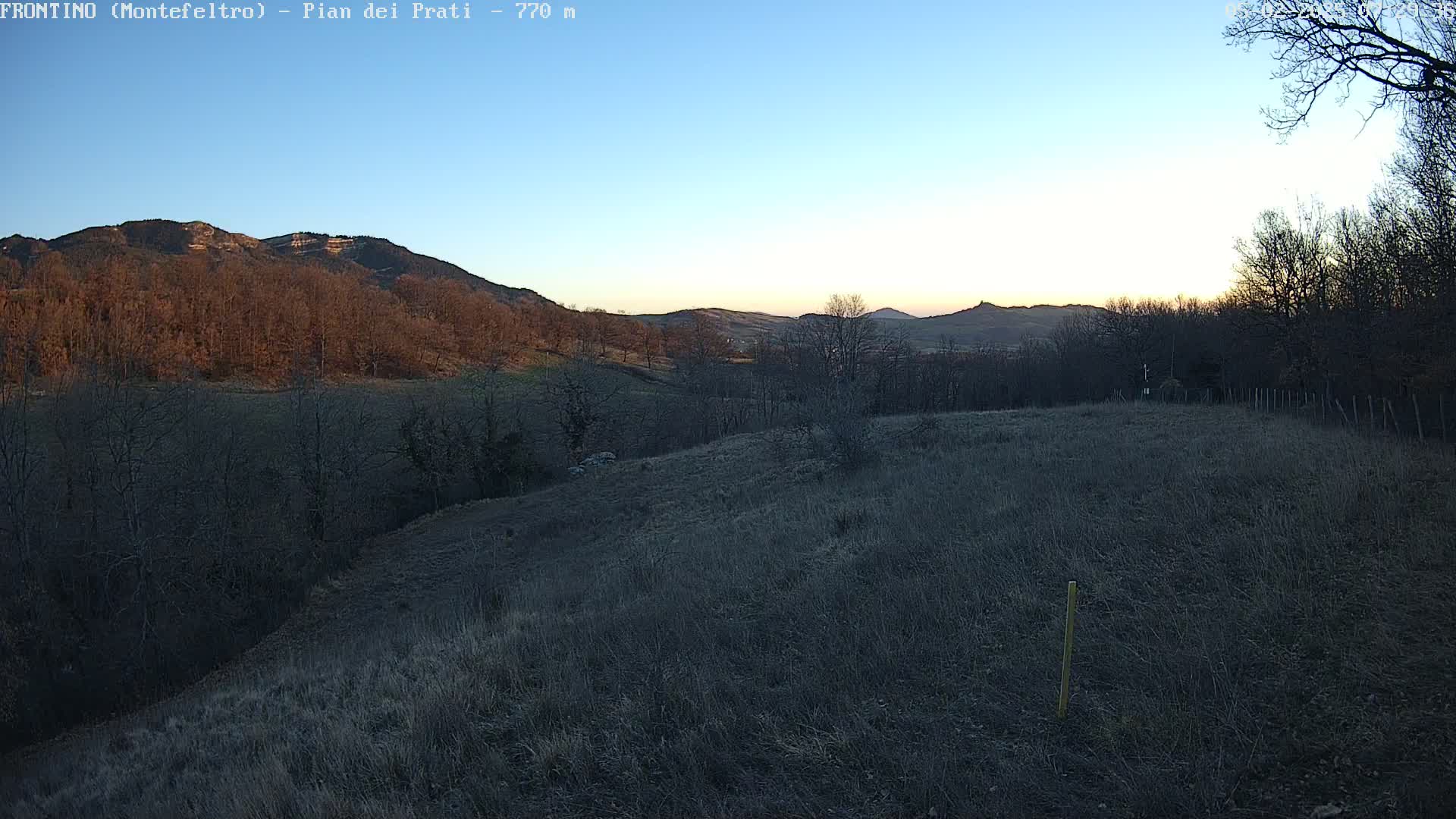 A view of a hilly landscape at sunset, showing a dry, grassy field in the foreground and bare, deciduous trees on the hillsides under a clear sky.