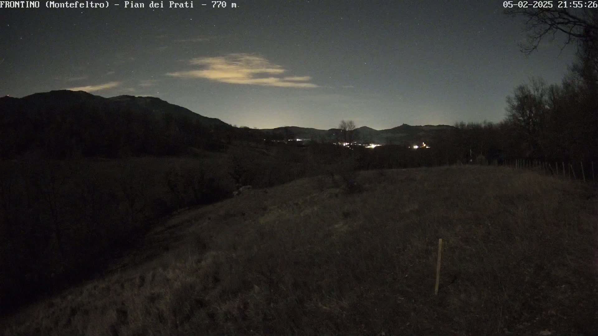 A nighttime, high-angle view shows a dark, grassy hillside under a mostly clear, starry sky with a few faint clouds.