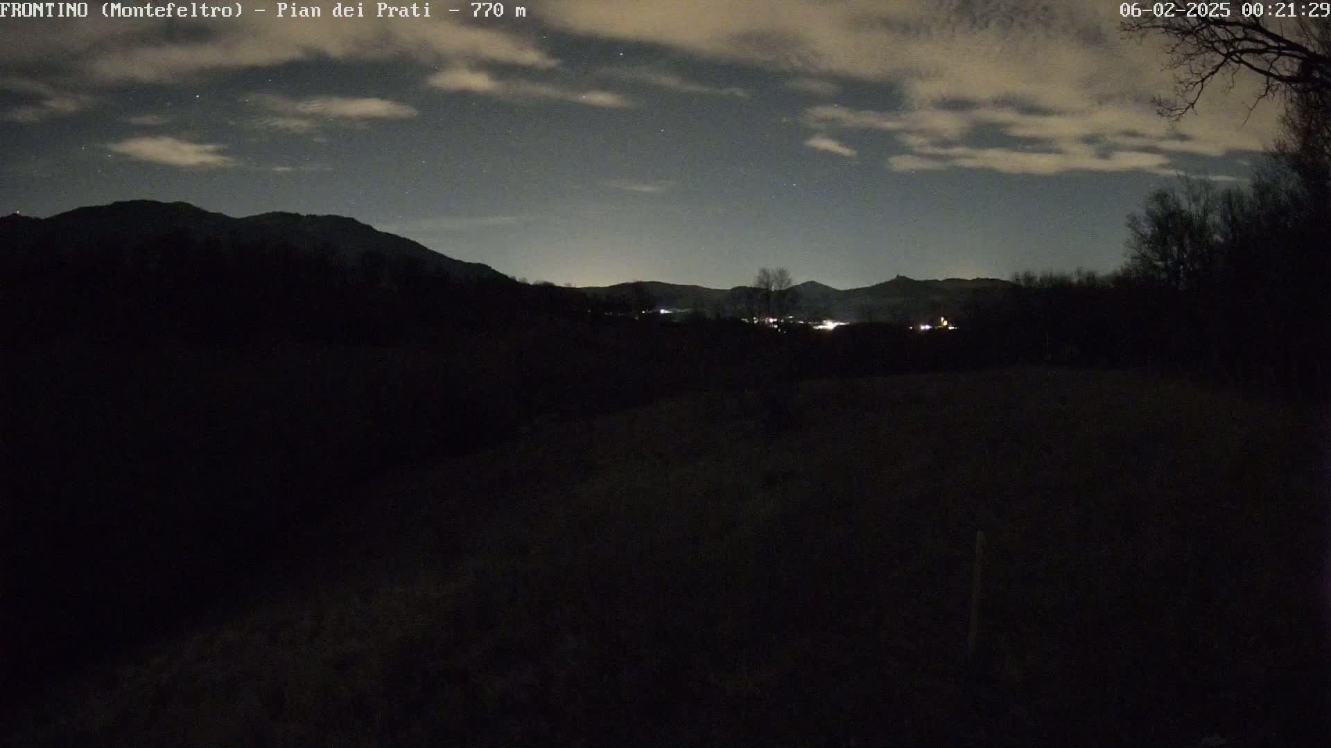 A nighttime landscape of dark, silhouetted mountains and trees under a partly cloudy sky with some stars visible.