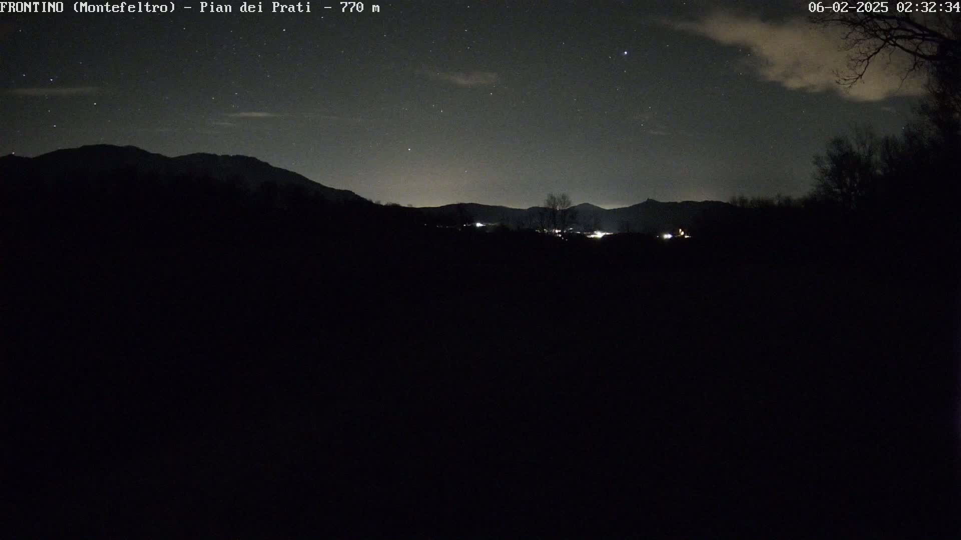 A nighttime view of a dark, mountainous landscape under a partly cloudy sky dotted with stars, showing some distant lights.