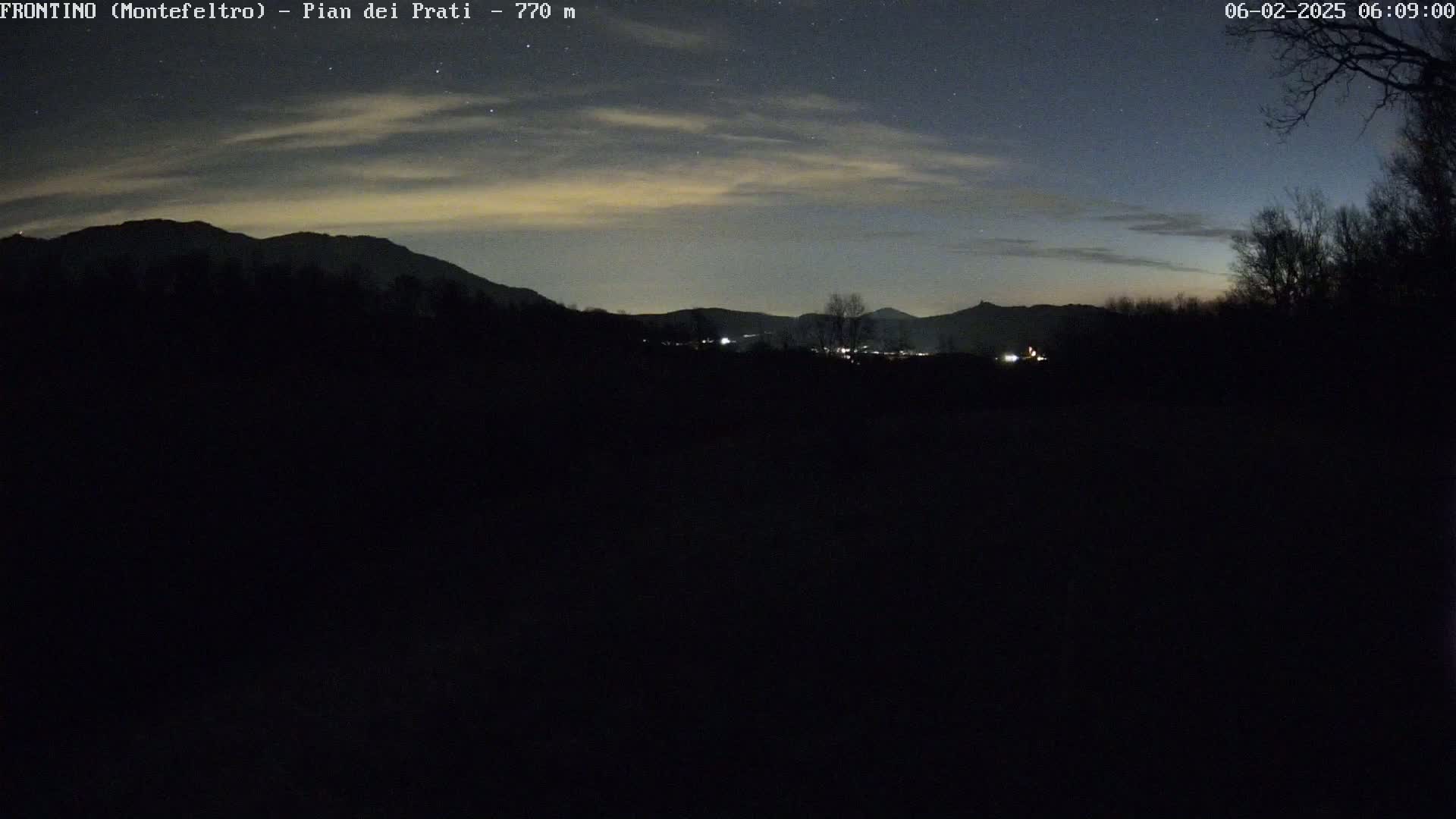 A nighttime view of a dark, silhouetted landscape featuring mountains under a mostly clear sky with some thin clouds and a few visible stars.