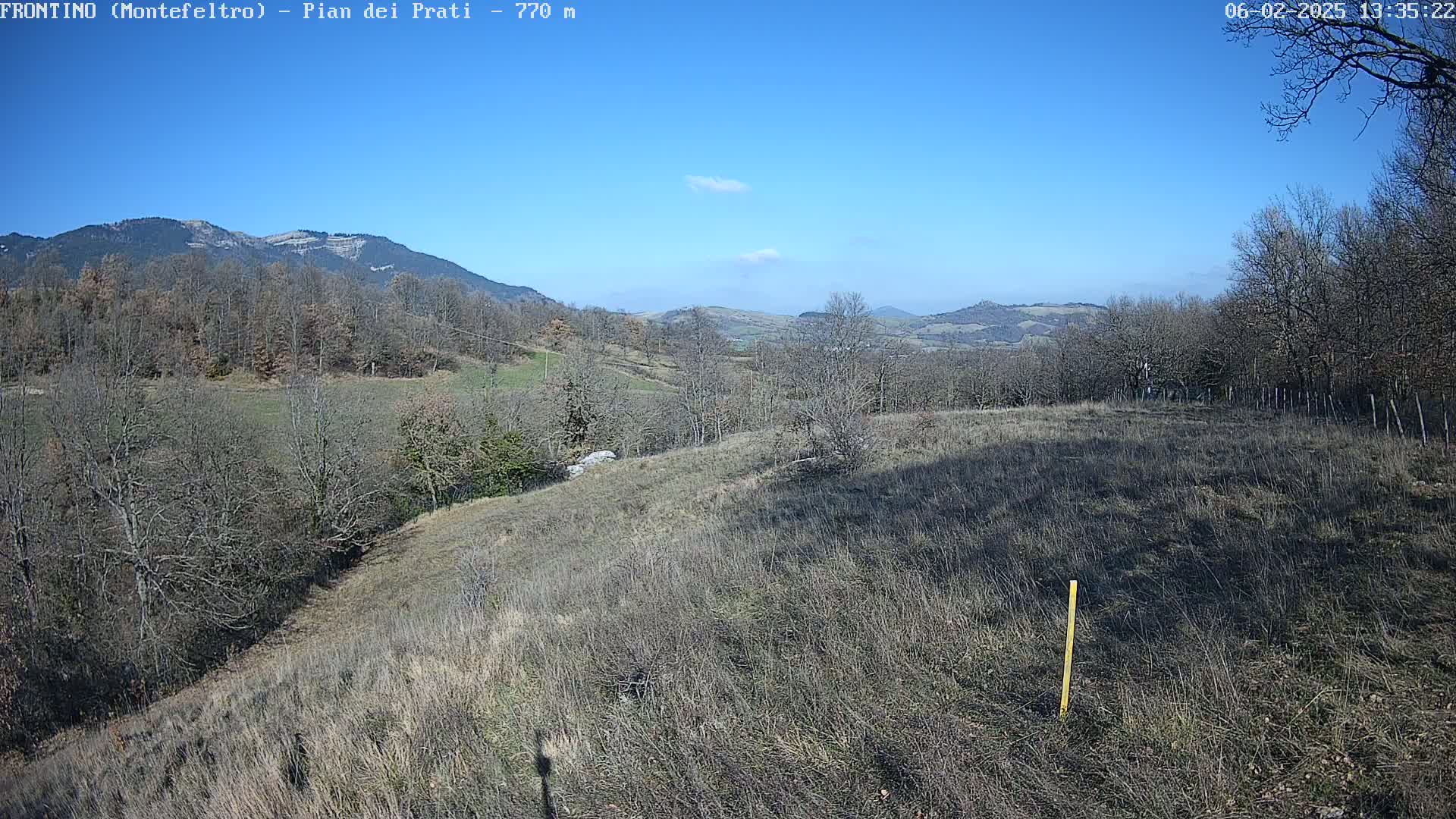 A mostly clear blue sky overlooks a hilly landscape of dormant, brown vegetation and leafless trees, under which a field stretches into the distance.