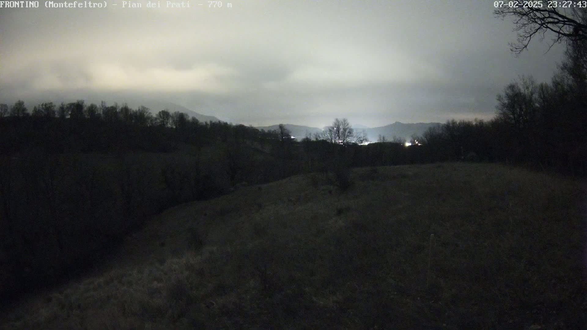 A dark, overcast night scene shows a hilly landscape with silhouetted trees and distant lights under a cloudy sky.