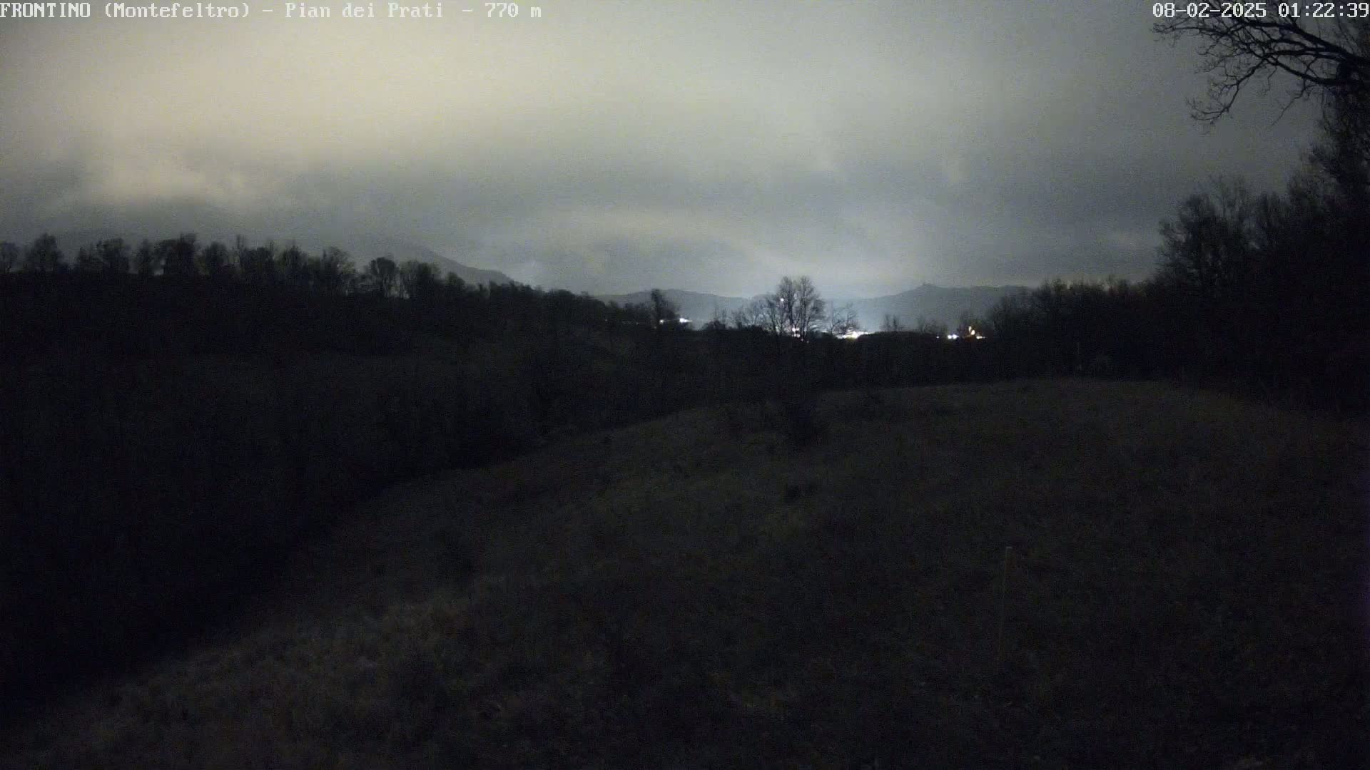 A nighttime view shows dark hills and trees under a cloudy, overcast sky, with faint lights visible in the distance.