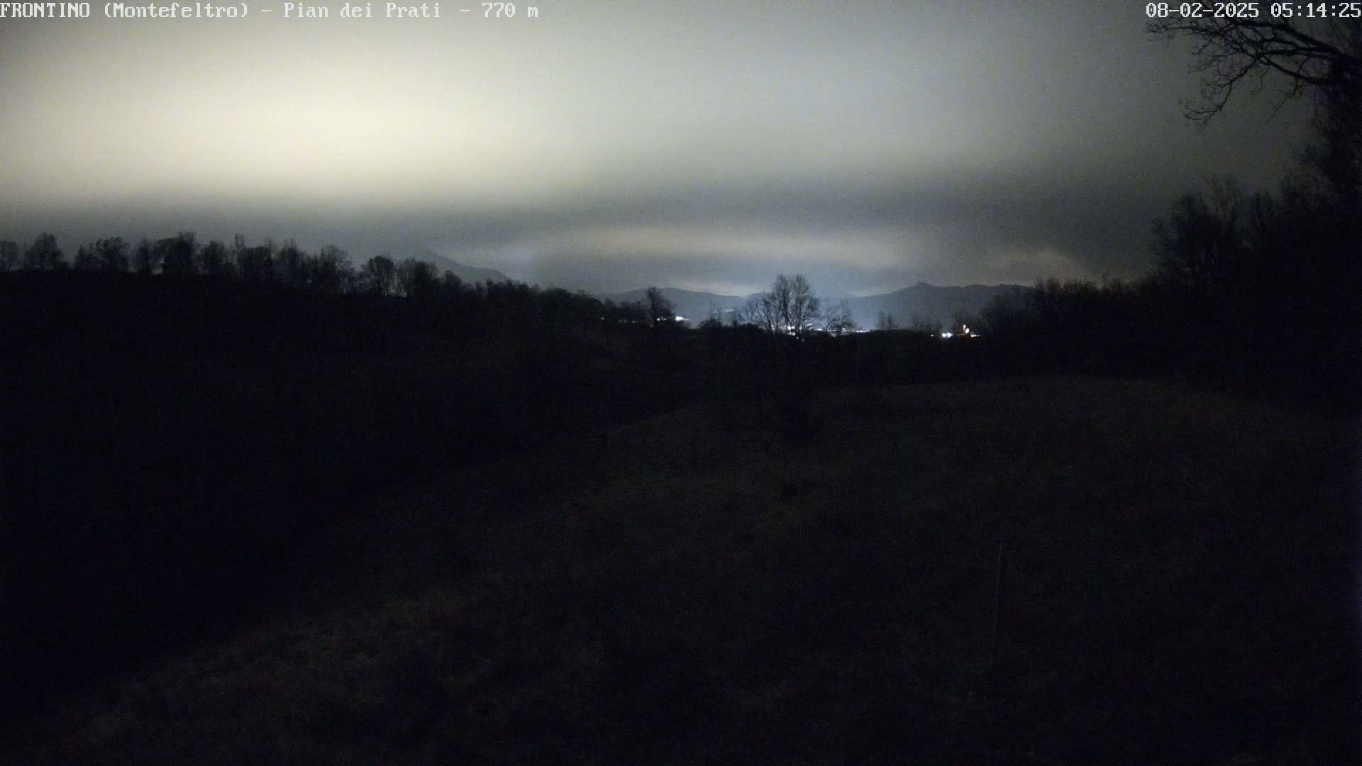 A dark, nighttime landscape shows silhouetted trees and distant mountains under a cloudy sky.