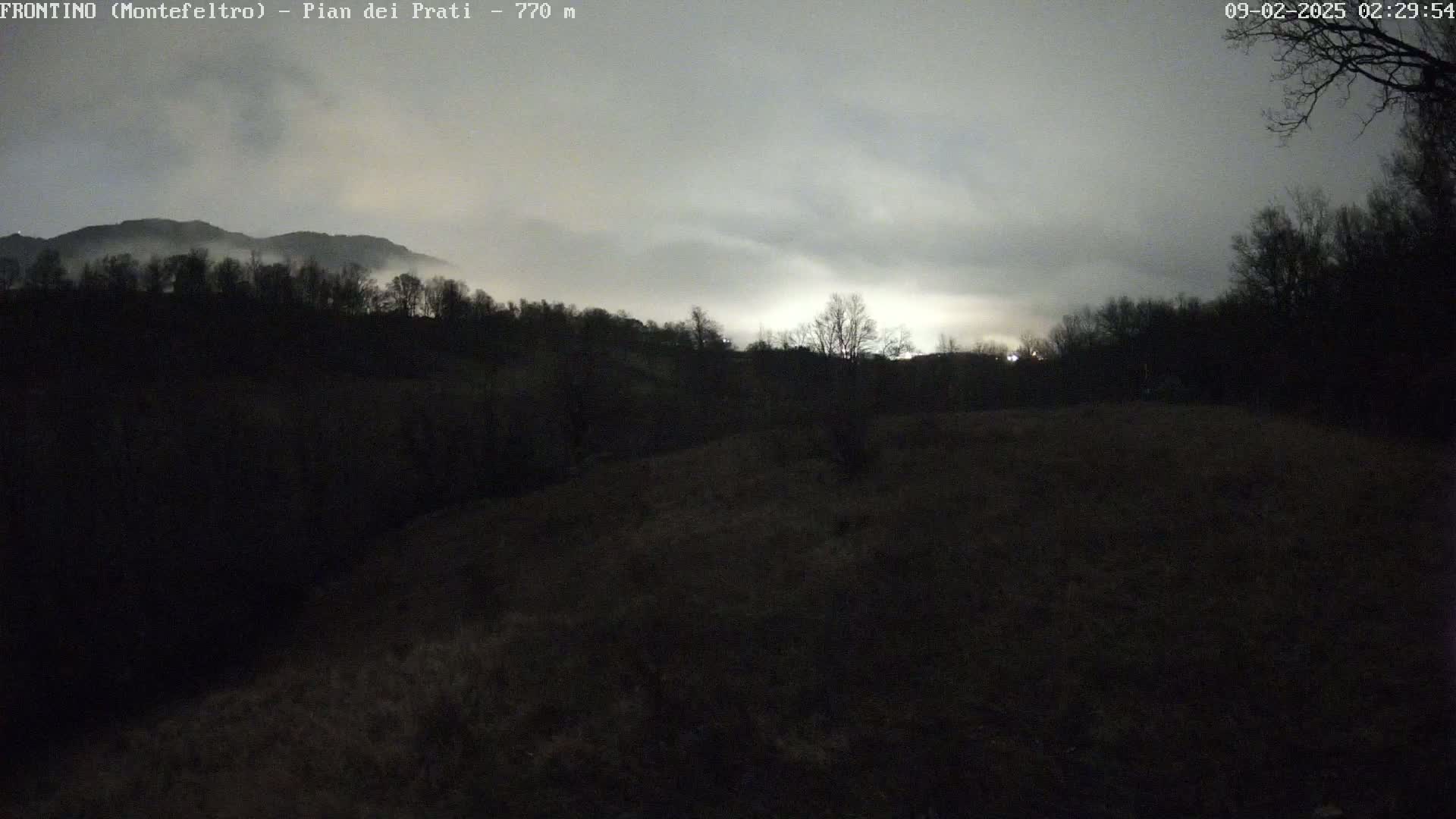 A dark, overcast night scene shows a hilly landscape with silhouetted trees and a distant, fog-shrouded mountain range.