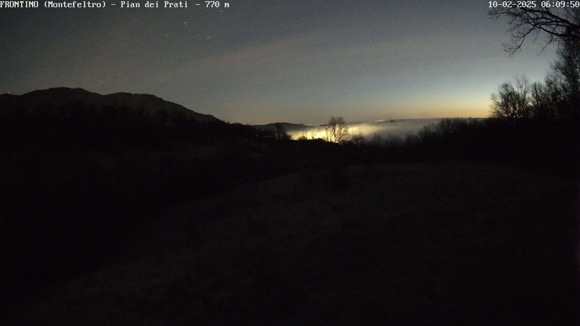 A low-lying fog covers a valley at dawn, illuminated by a few distant lights, under a dark, starry sky.