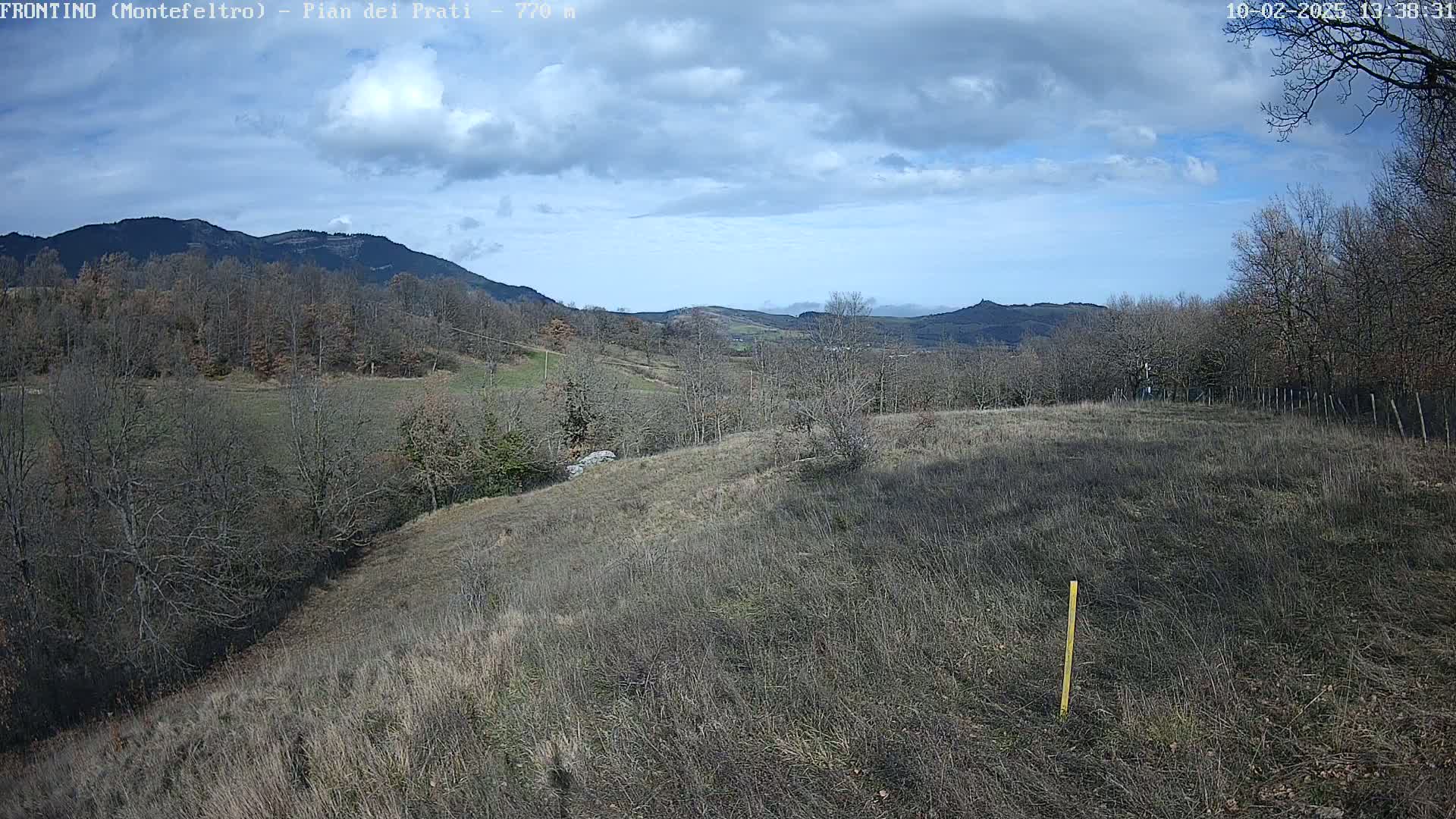 A partly cloudy day reveals a hilly landscape with leafless trees and brown, grassy fields, extending to distant mountains under a mostly clear sky.