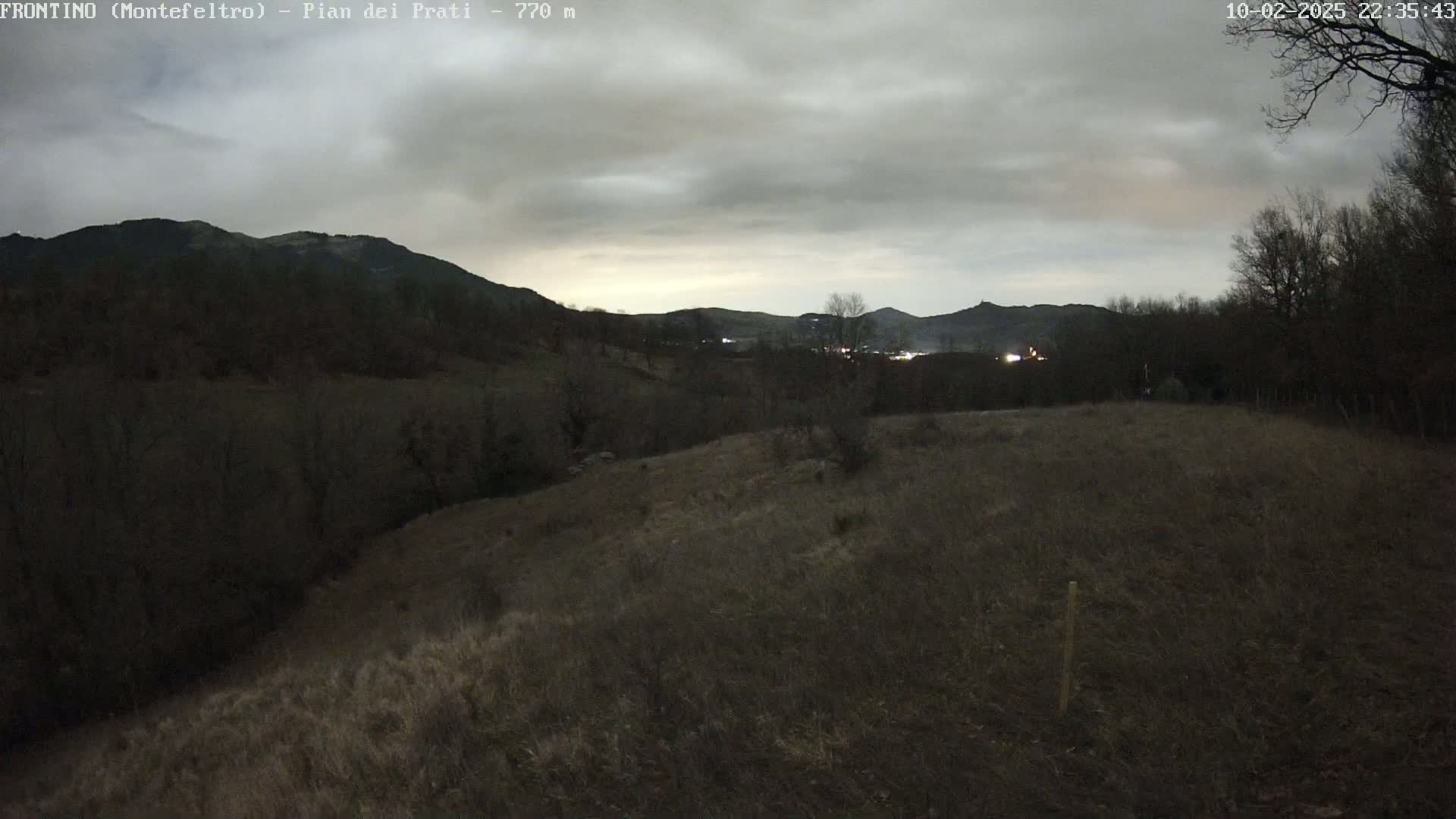 A view from a hilltop shows a valley with dark hills and a few lights in the distance under an overcast sky.