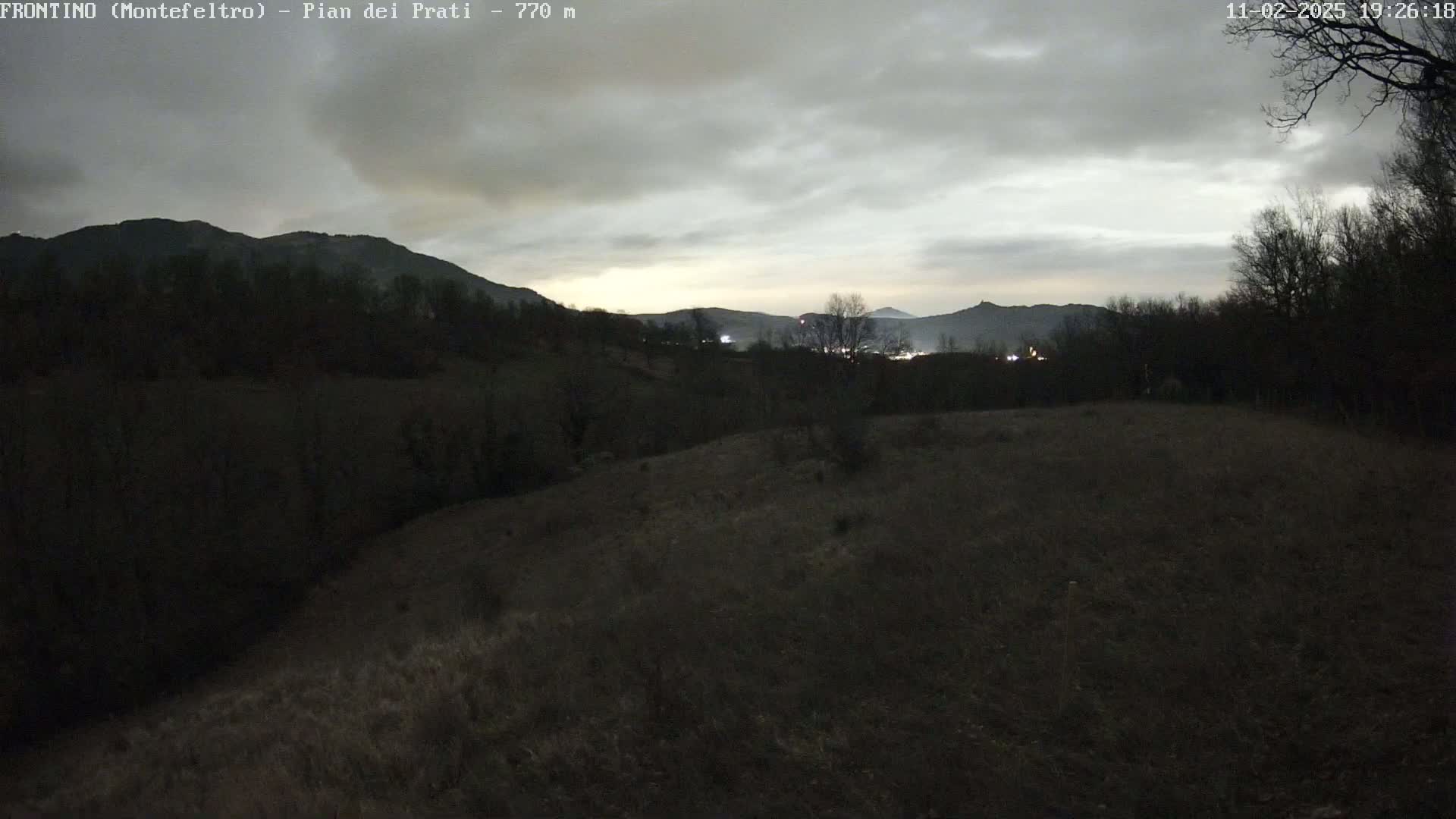 A dimly lit, overcast landscape at dusk shows dark, silhouetted hills and trees, with a field in the foreground.