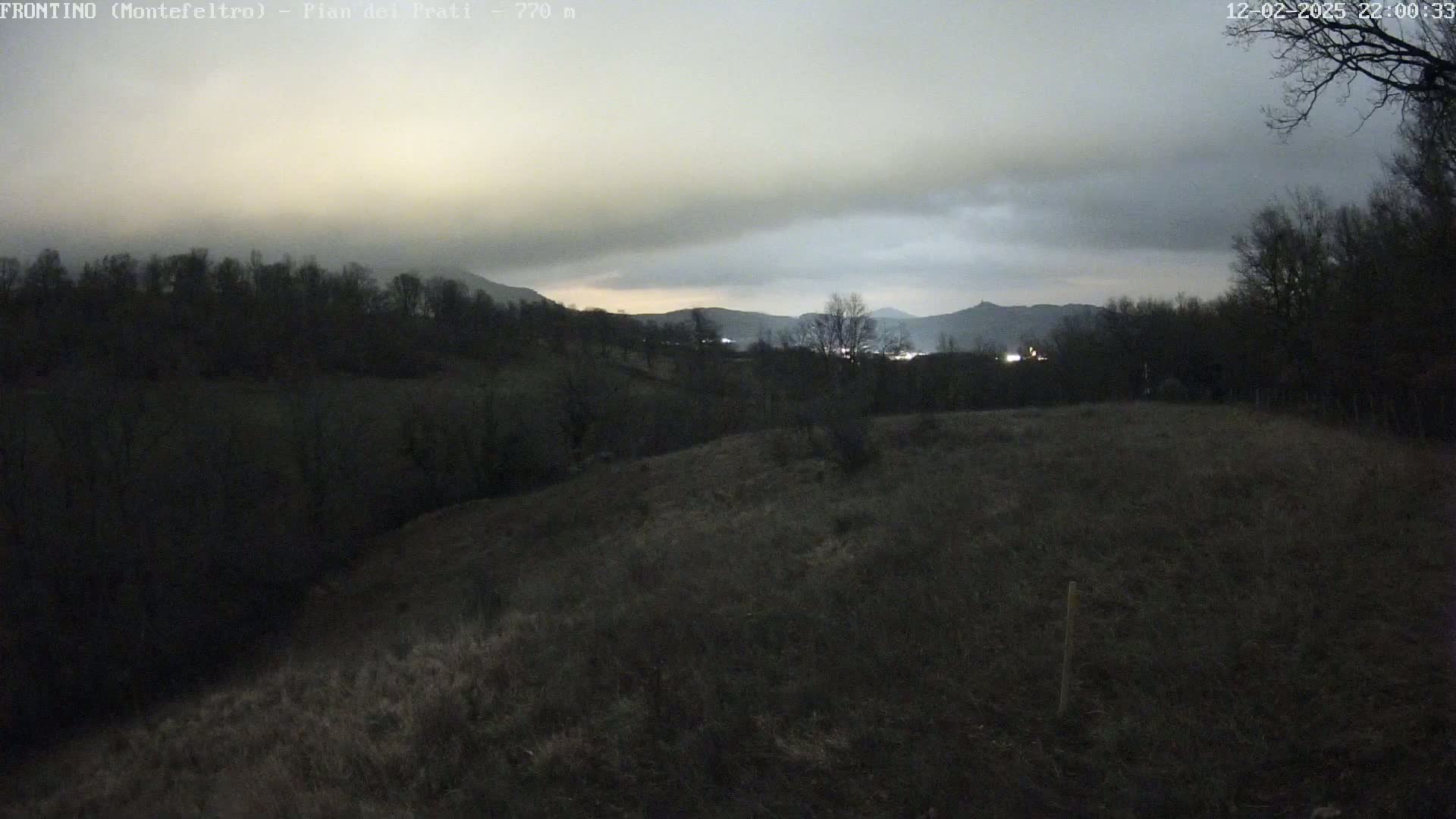A dark, overcast evening scene shows a hilly landscape with sparse vegetation, trees lining the horizon, and distant lights visible under a cloudy sky.
