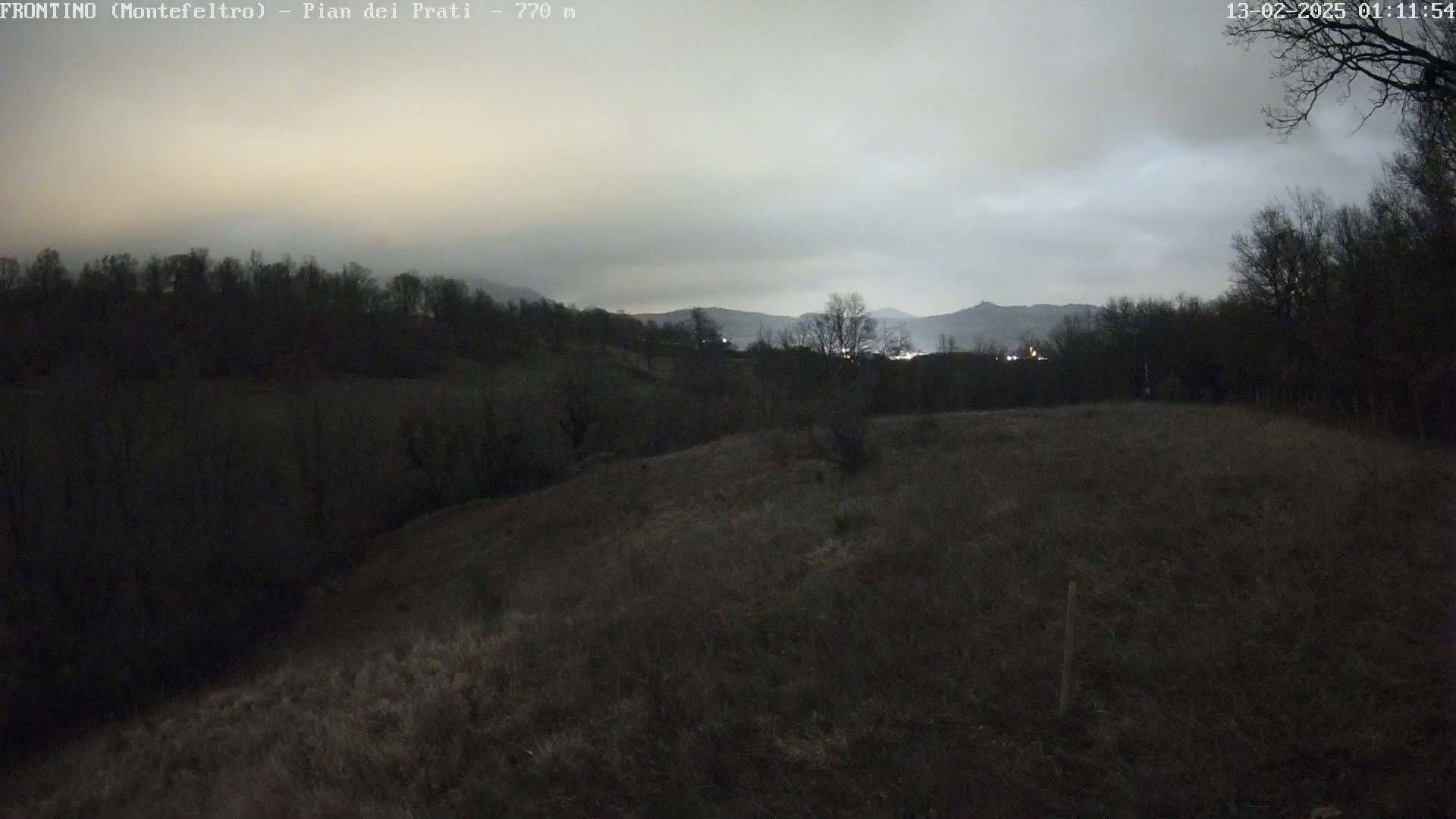 A dark, overcast evening scene shows a hilly landscape with bare trees and fields under a cloudy sky.