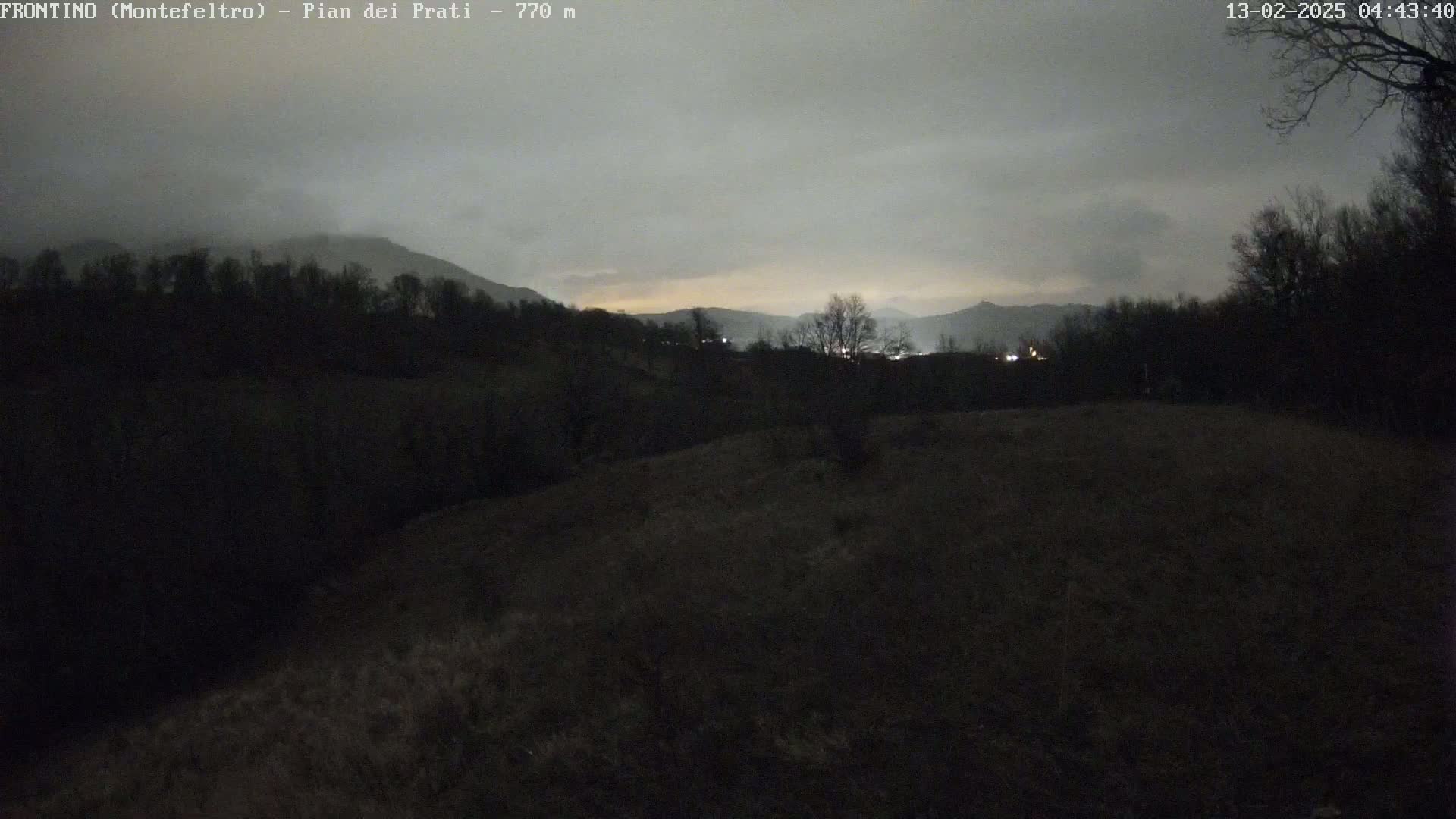 A dark, overcast night scene shows a hilly landscape with sparse trees and a dimly lit distant town under a cloudy sky.