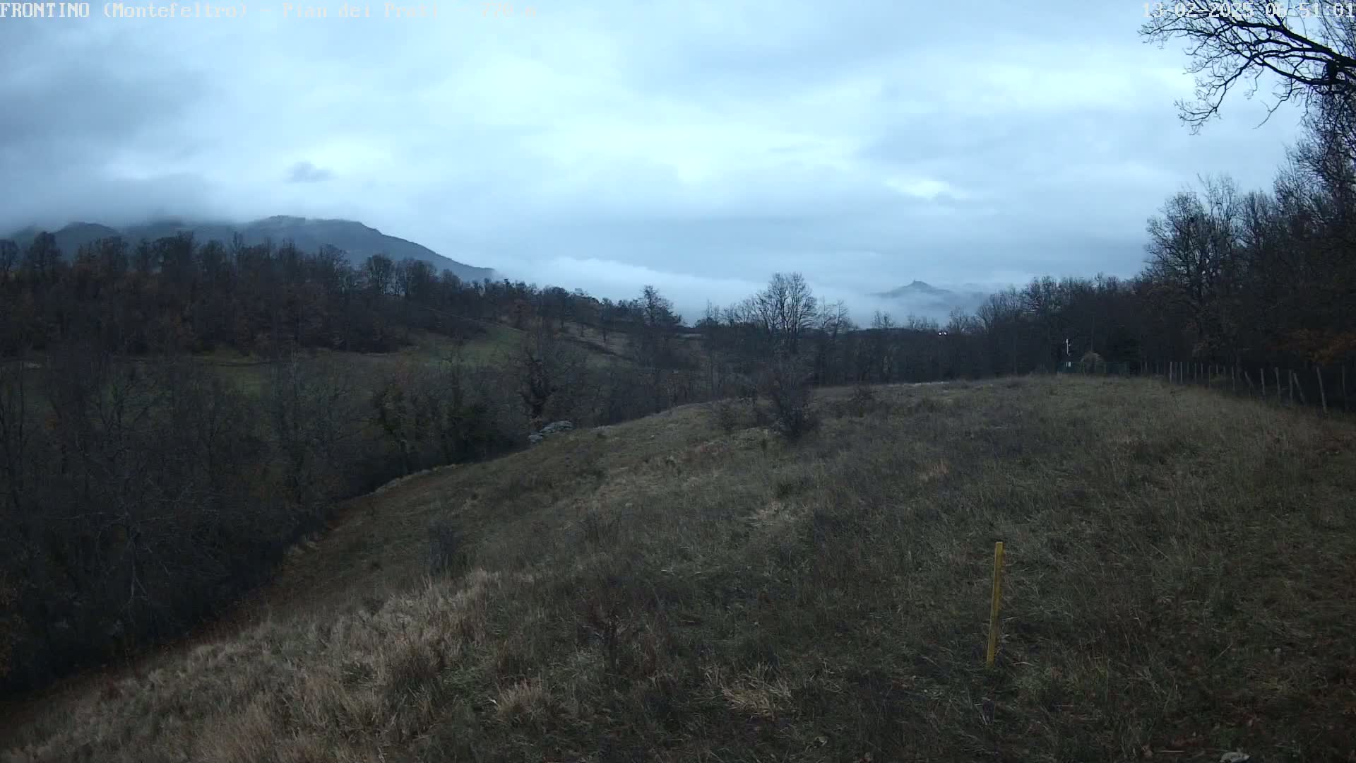 A hilly landscape with leafless trees under a cloudy, overcast sky shows a valley fog partially obscuring distant mountains.