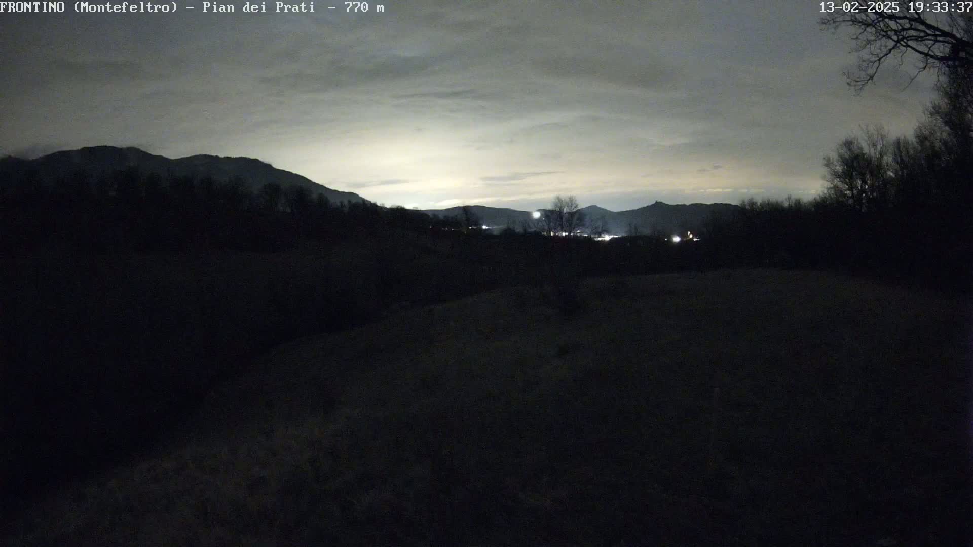 A nighttime view of dark, silhouetted mountains and trees under a cloudy, light gray sky.