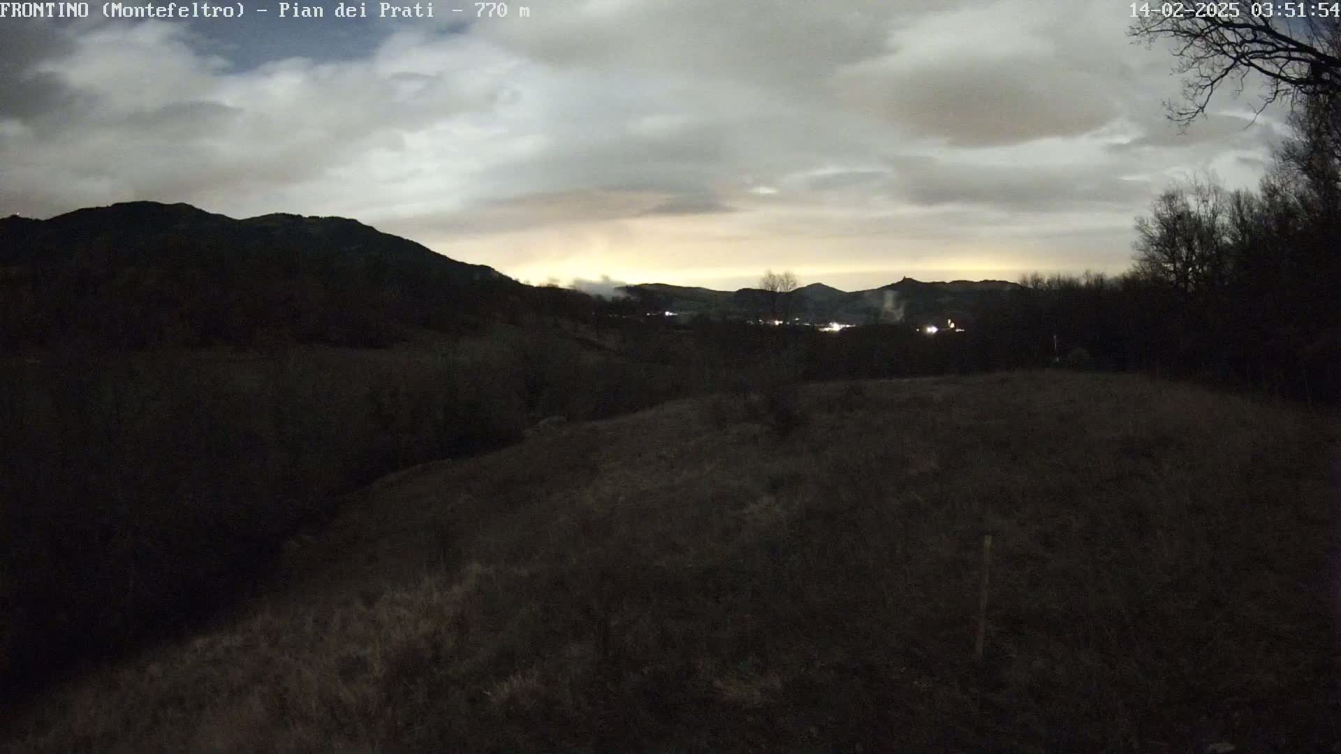 A dark, overcast evening scene shows a rolling landscape of hills and sparse vegetation under a cloudy sky, with a hint of light on the horizon.