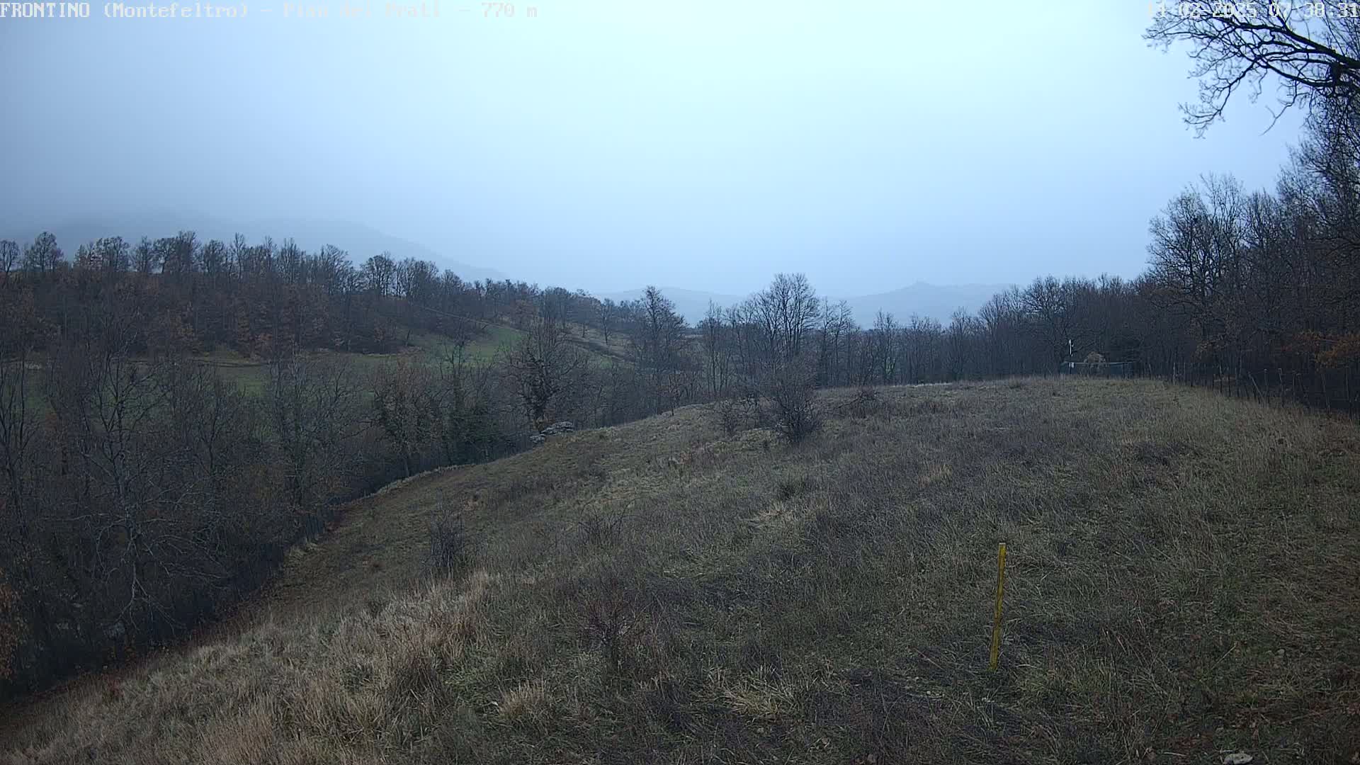 A hazy, overcast day reveals a rolling landscape of barren, brown fields and leafless trees, with distant mountains barely visible in the muted light.