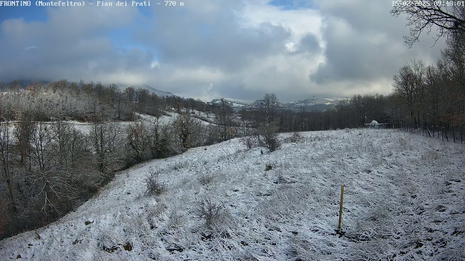 A snow-covered field and hillside with leafless trees under a cloudy sky.