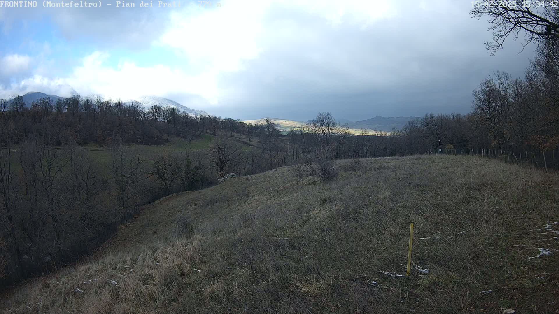 A partly cloudy day reveals a hilly landscape with bare, winter trees and a snow-capped mountain in the distance.
