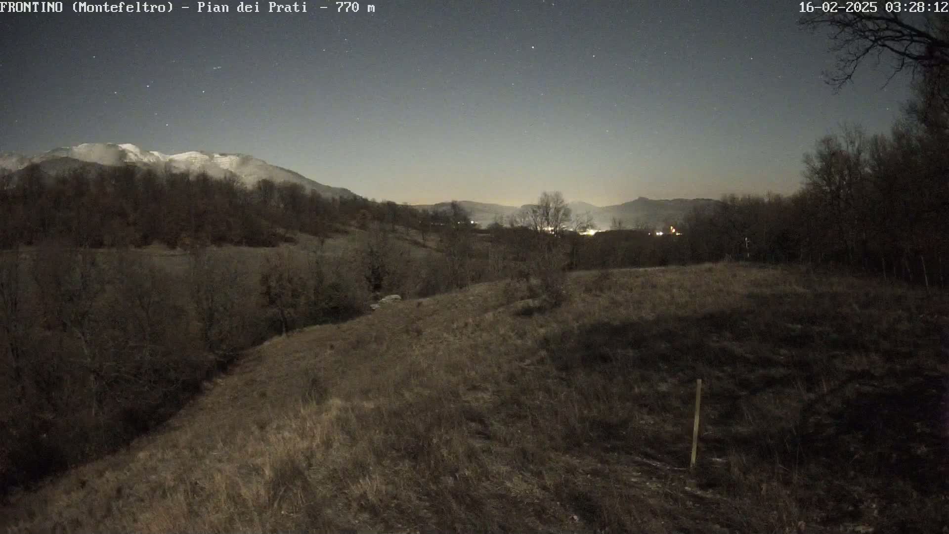 A snow-capped mountain is visible in the distance under a clear, starry night sky, overlooking a dry, hilly landscape with sparse vegetation.