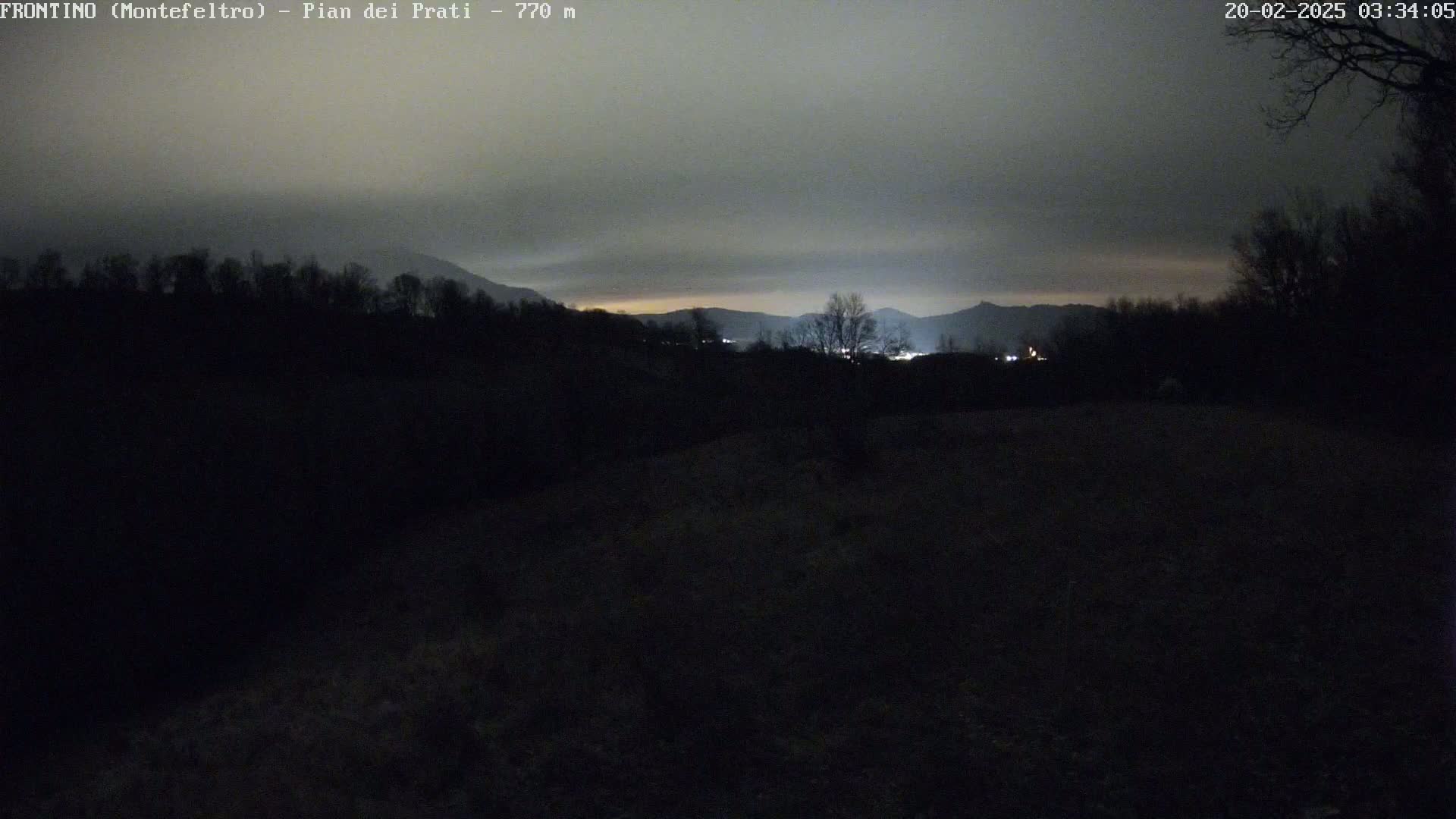 A nighttime, overcast view shows dark, silhouetted trees and hills, with faint lights visible in the distance.