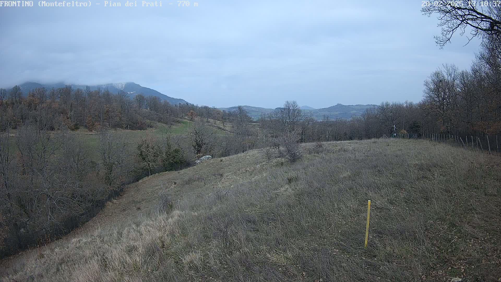A mostly cloudy day reveals a hilly landscape with bare trees and dry, brown grass, distant mountains with a hint of snow on their peaks.