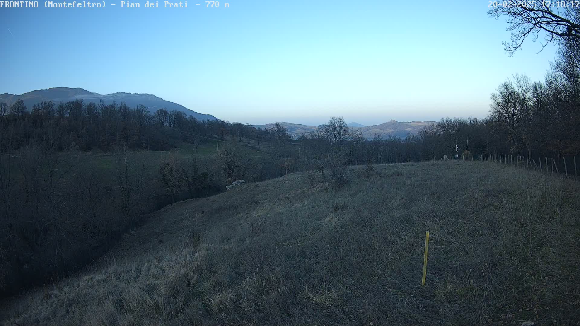A sprawling, dry field slopes gently downward towards a line of leafless trees and distant mountains under a clear, light blue sky.