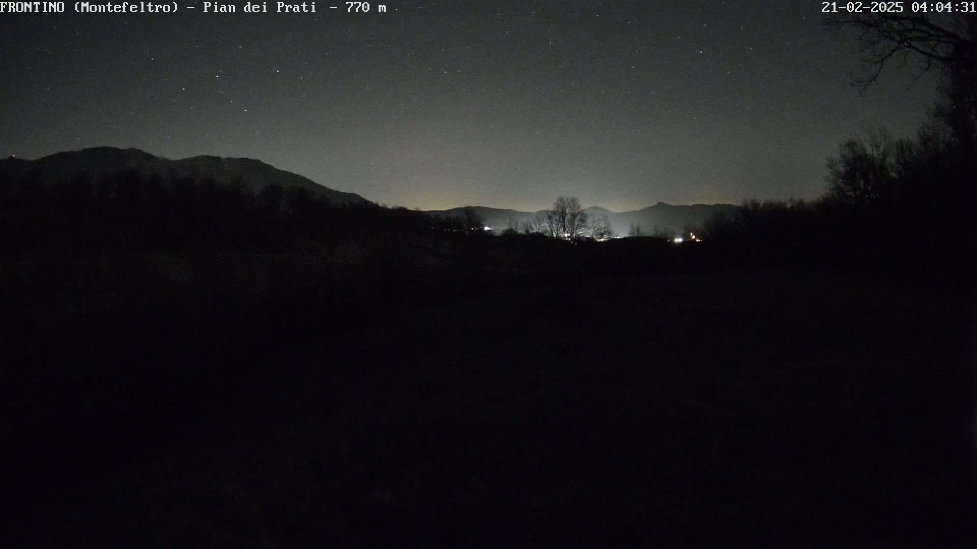 A nighttime, starlit view of dark silhouettes of mountains and trees, with a few faint lights visible in the distance.