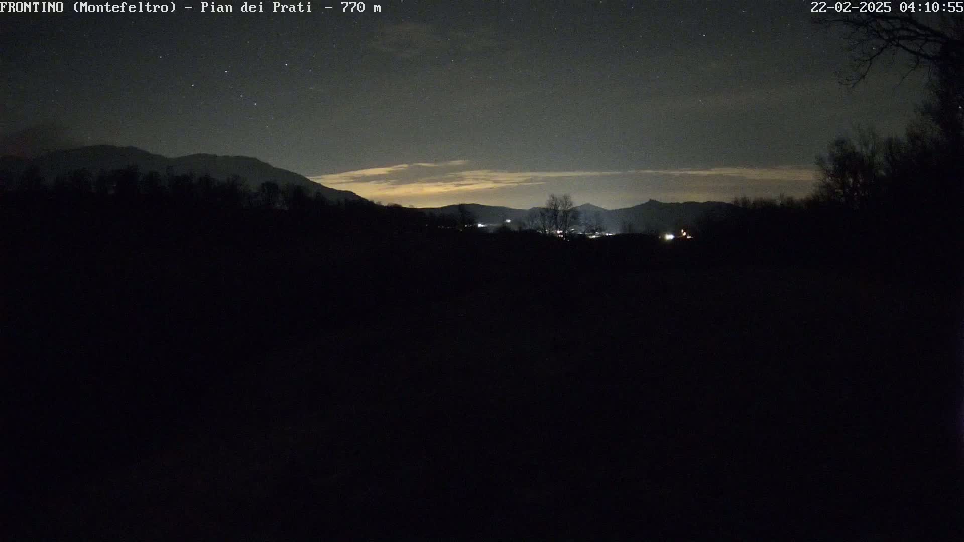A nighttime view of silhouetted mountains and trees under a dark, starry sky with a faint, light orange horizon.