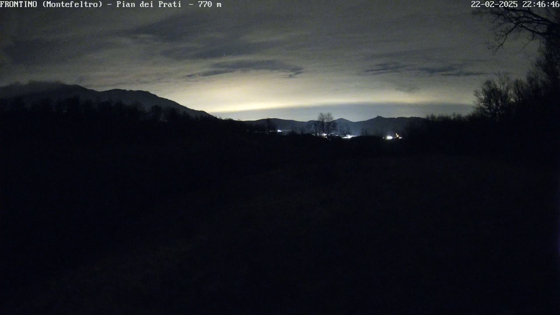 A nighttime view of silhouetted mountains and trees under a cloudy sky with a faint, yellowish glow on the horizon.