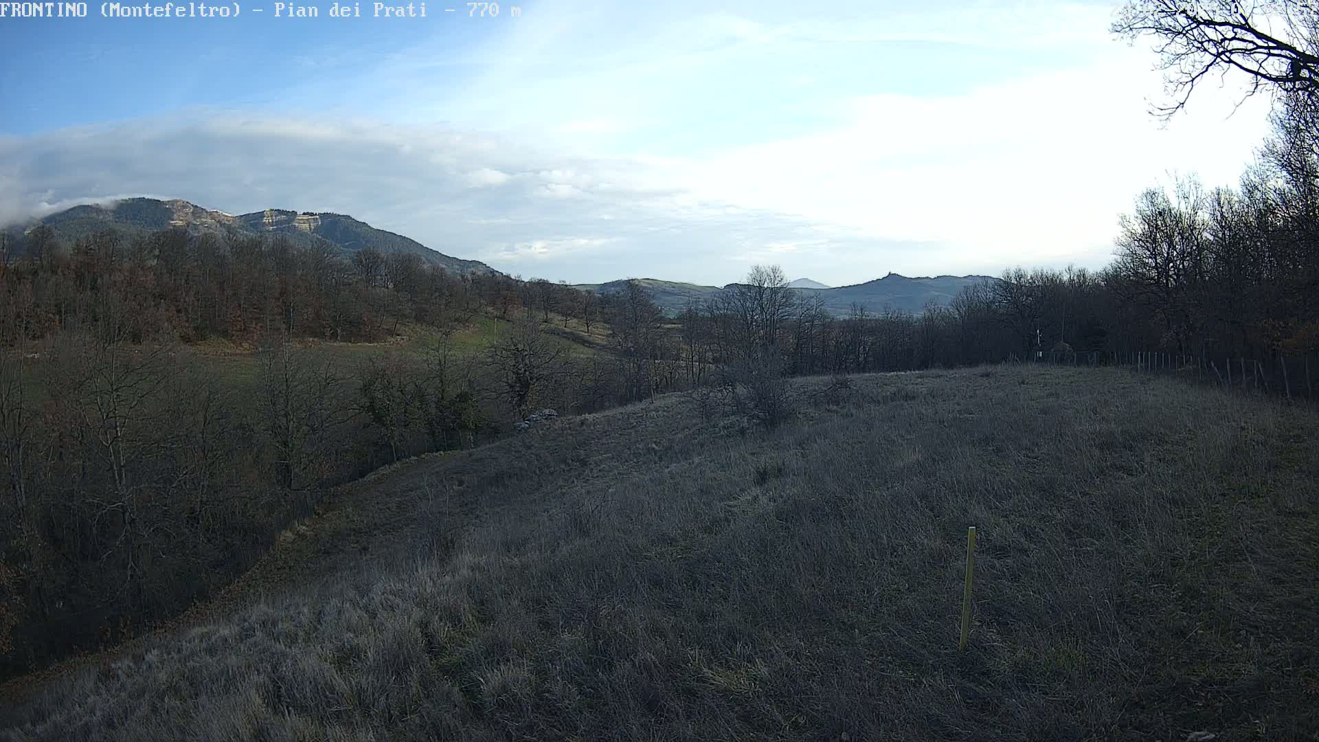 A mostly barren, hilly landscape with leafless trees and distant mountains under a partly cloudy sky.
