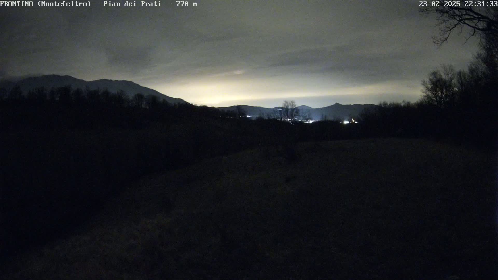 A nighttime view of dark, silhouetted trees and hills under a cloudy sky, with a faint, yellowish glow on the horizon.