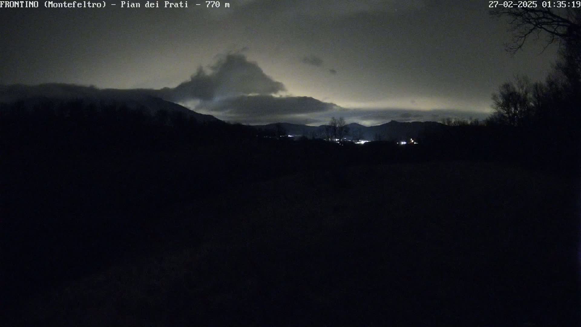 A nighttime view of dark, silhouetted mountains and trees under a cloudy sky with faint light sources visible in the distance.