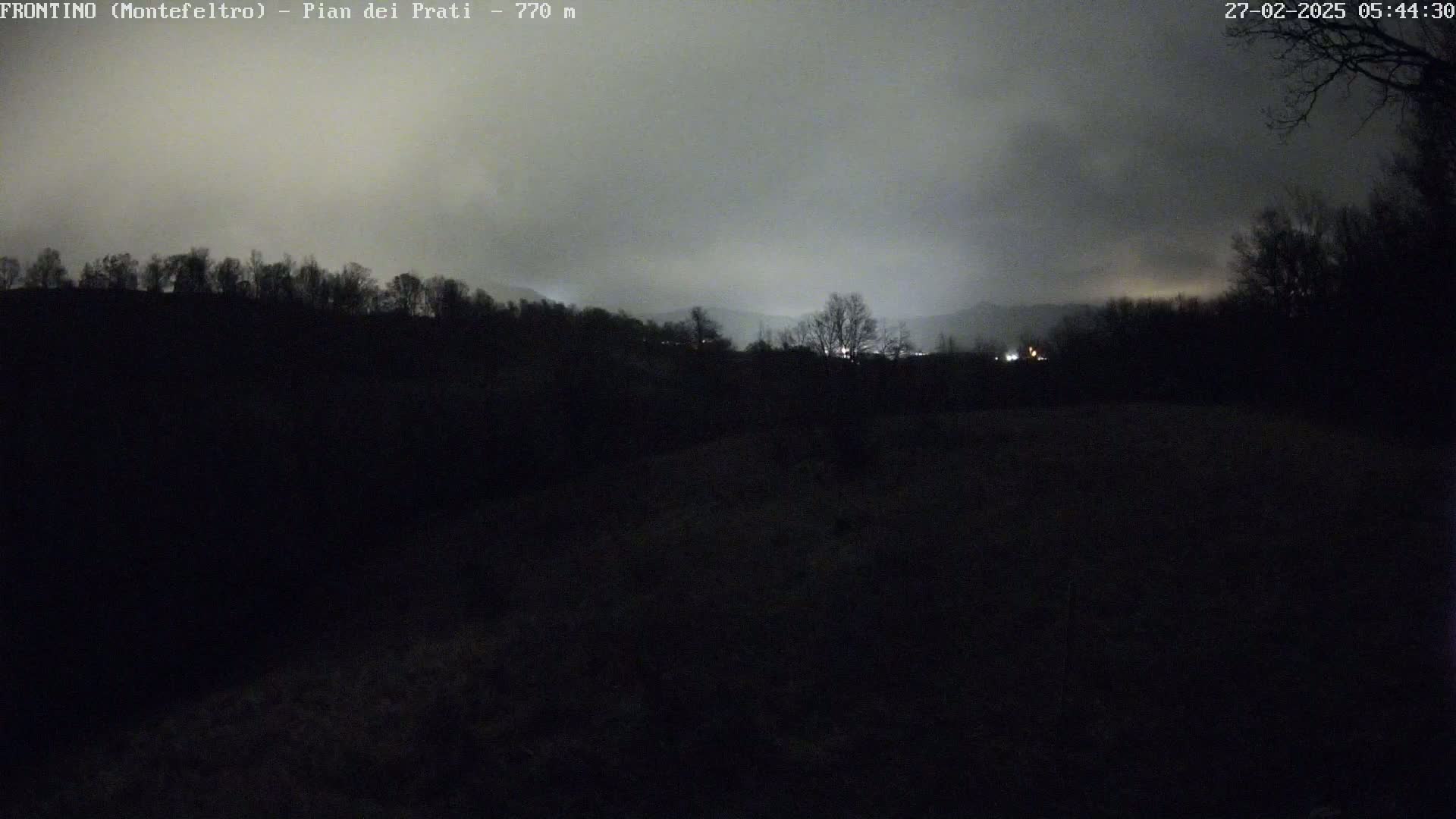 A dark, overcast night scene shows a silhouetted landscape of trees and a field, with faint lights visible in the distance.