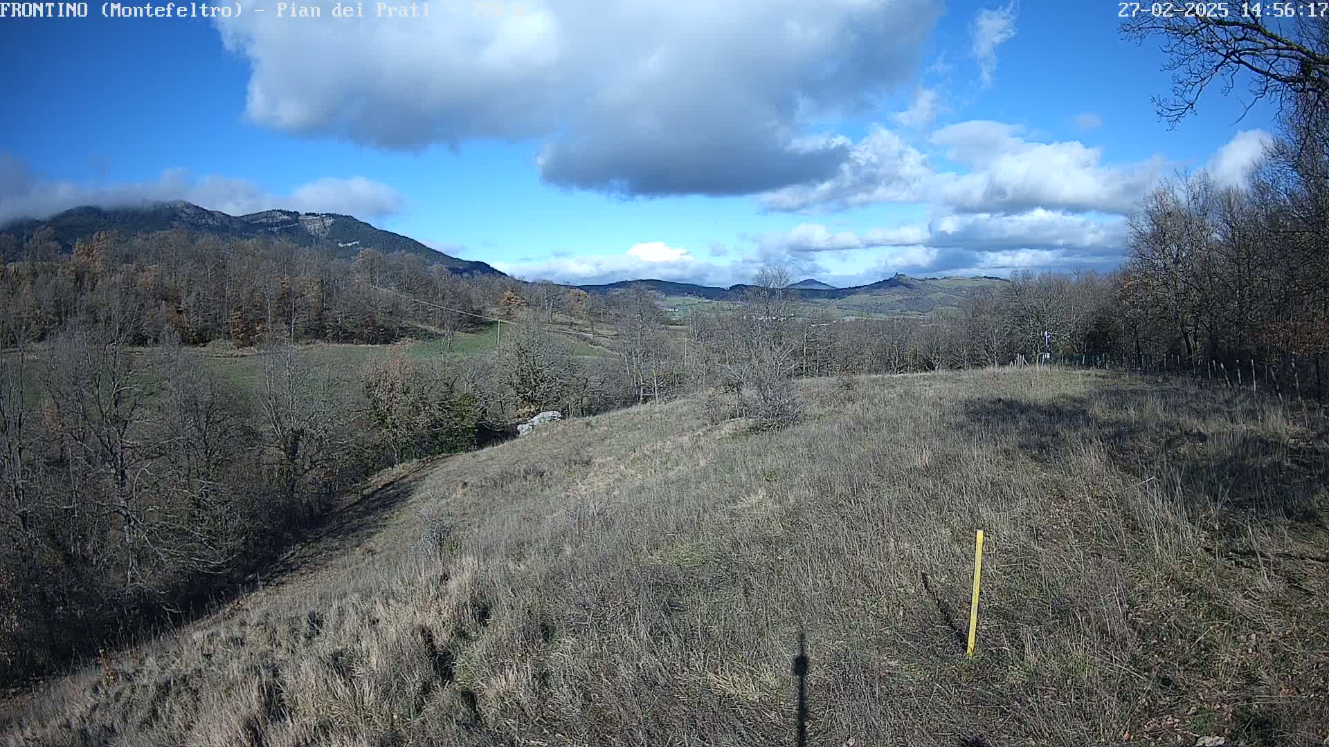 A partly cloudy blue sky overlooks a hilly landscape of brown, dry grass and leafless trees, with distant mountains in the background.