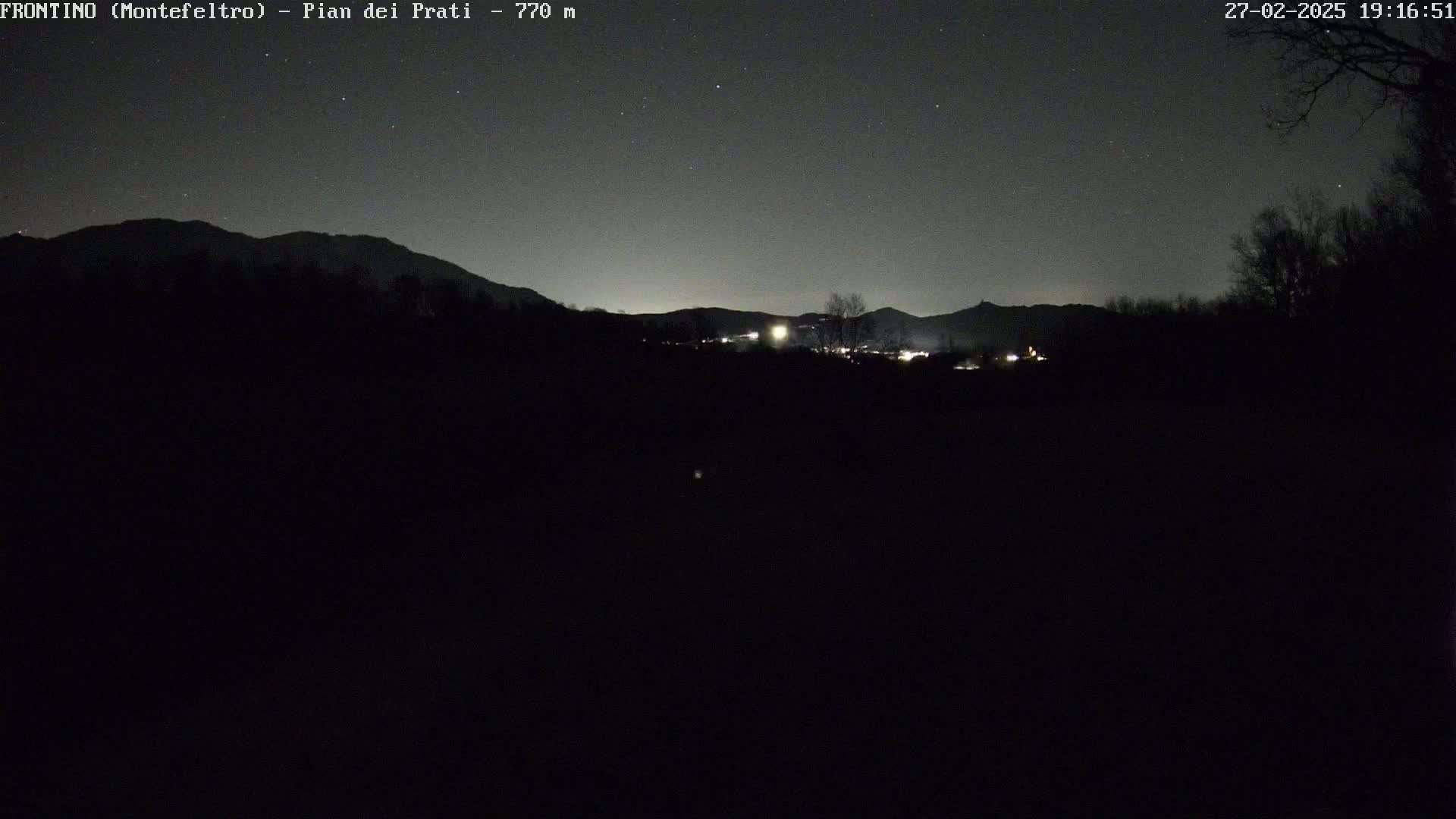 A nighttime view of dark silhouettes of distant mountains and trees under a starry sky, with a few faint lights visible in the middle distance.