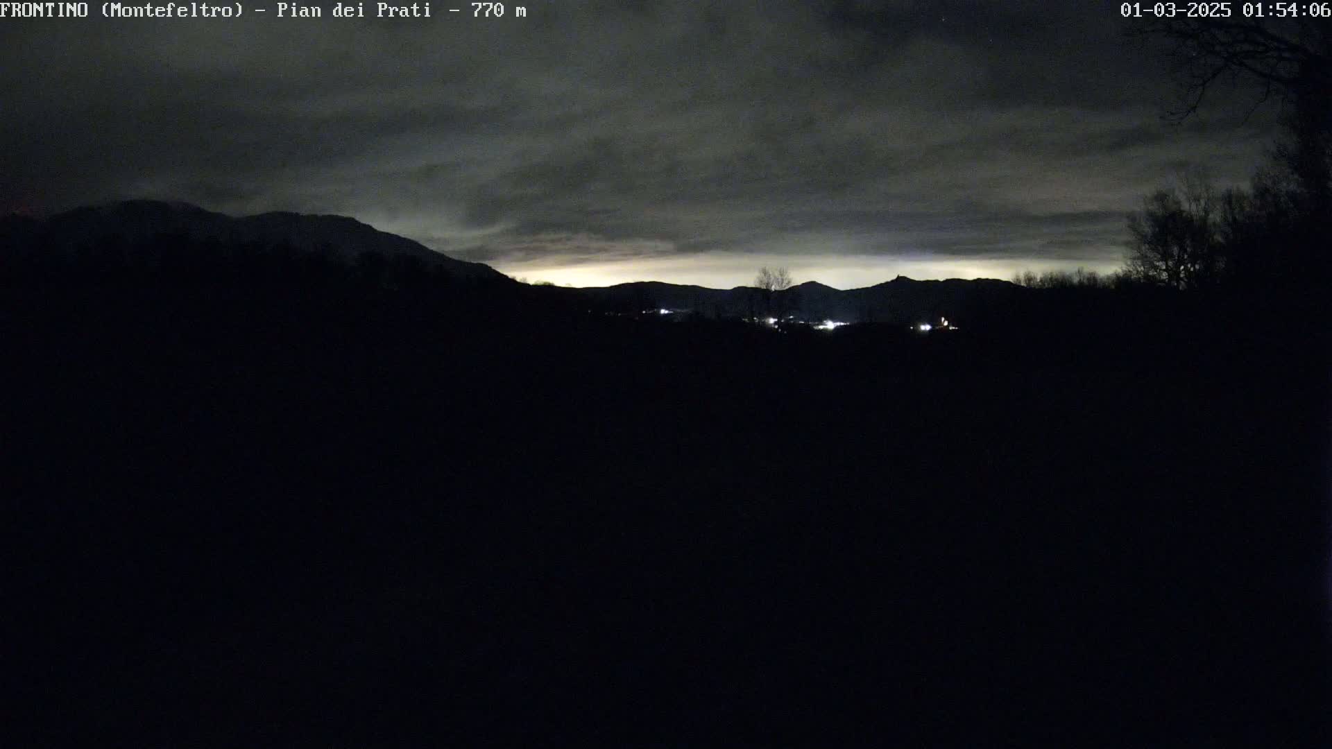 A dark, nighttime landscape shows silhouetted mountains under a cloudy sky, with a few faint lights visible in the distance.