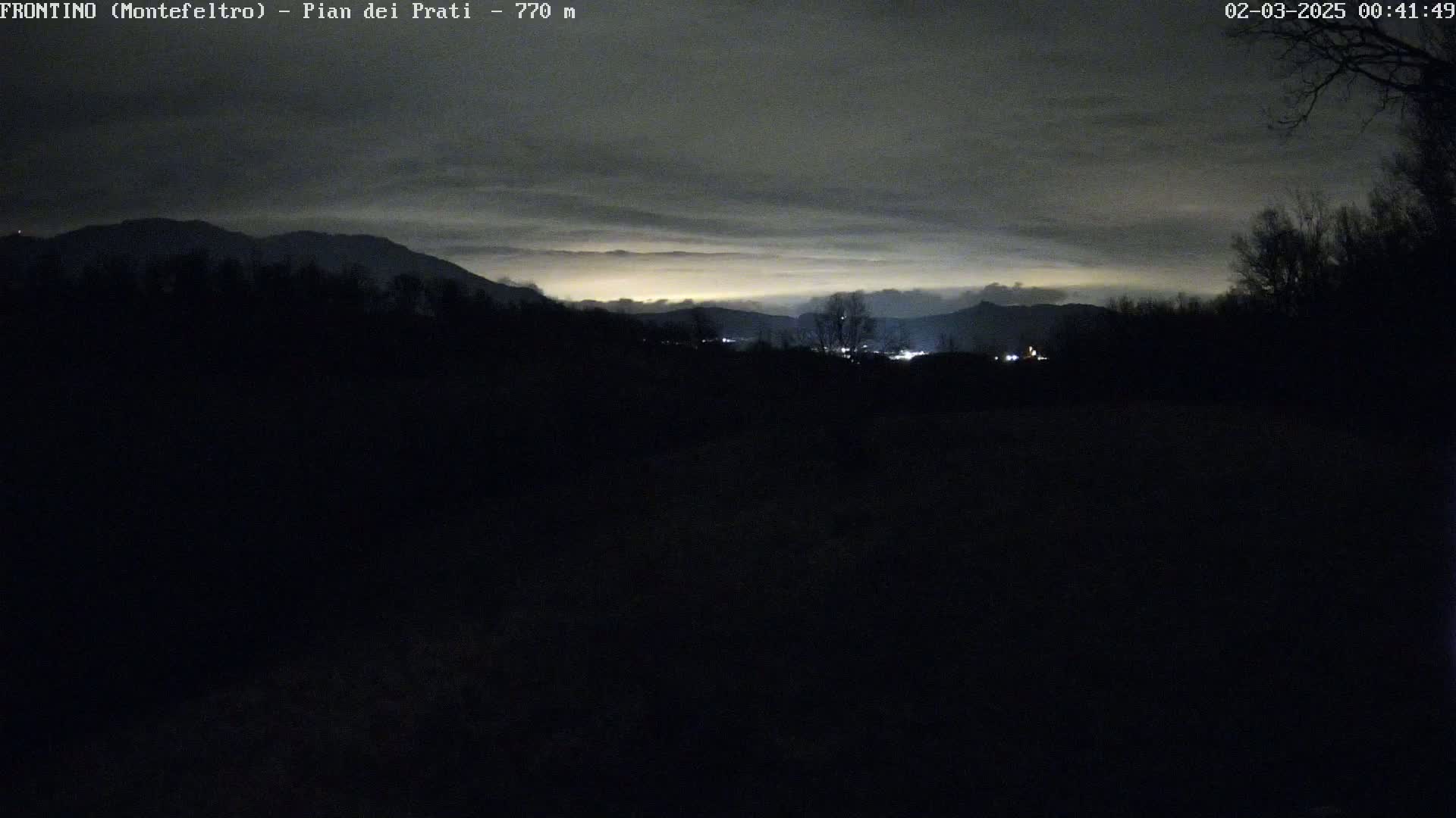 A nighttime view of a dark, hilly landscape with a faintly lit town in the distance under a cloudy sky.